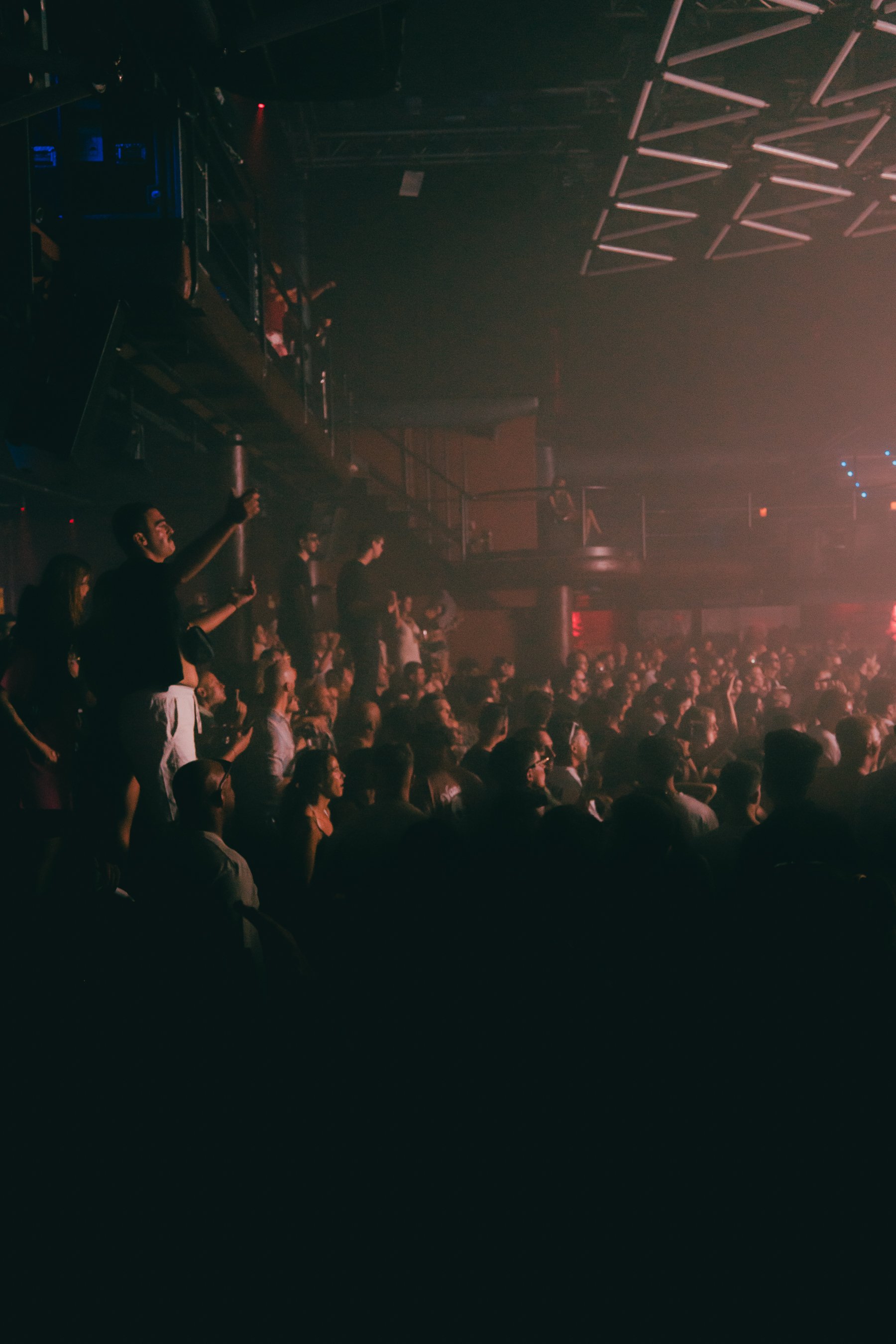 A large crowd of people gathered in an indoor concert or event space, with some standing and some sitting, under a ceiling with geometric light fixtures, dim lighting, and a stage or performance area not visible in the image.