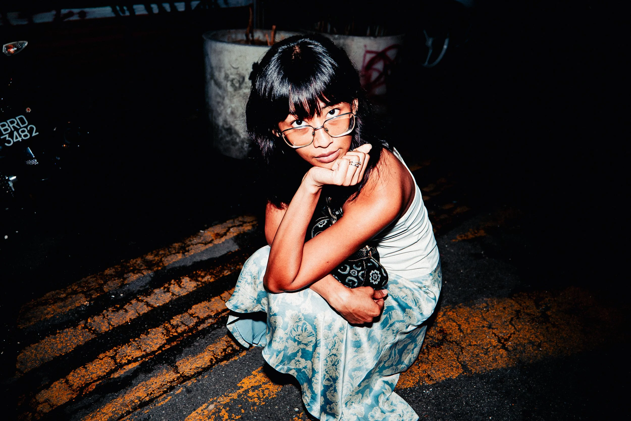 A woman with glasses and dark hair sits on a curb at night, resting her chin on her hand, with a potted plant and a motorcycle license plate visible behind her.