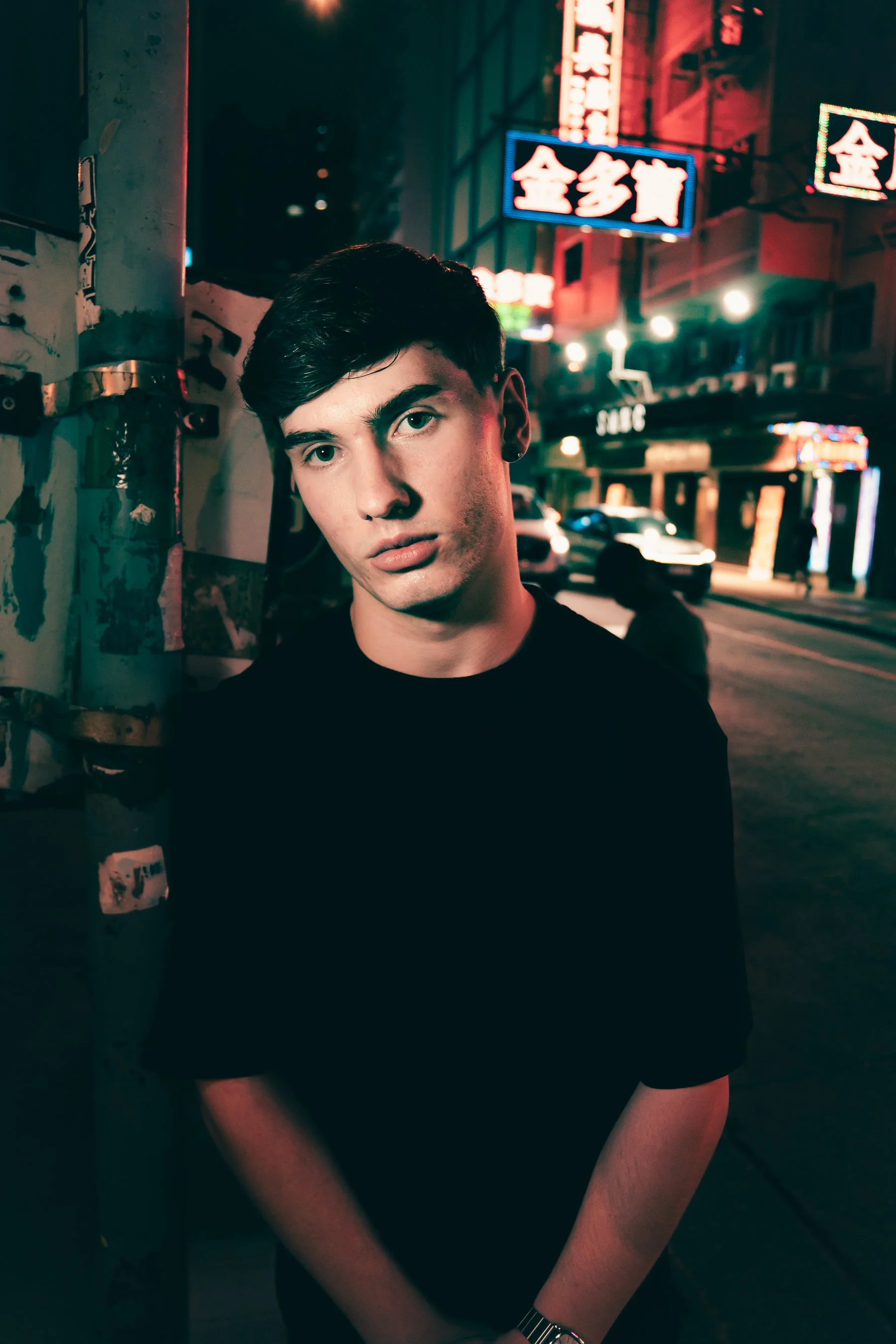 A young man with dark hair and a serious expression standing on a city street at night with neon signs and cars in the background.