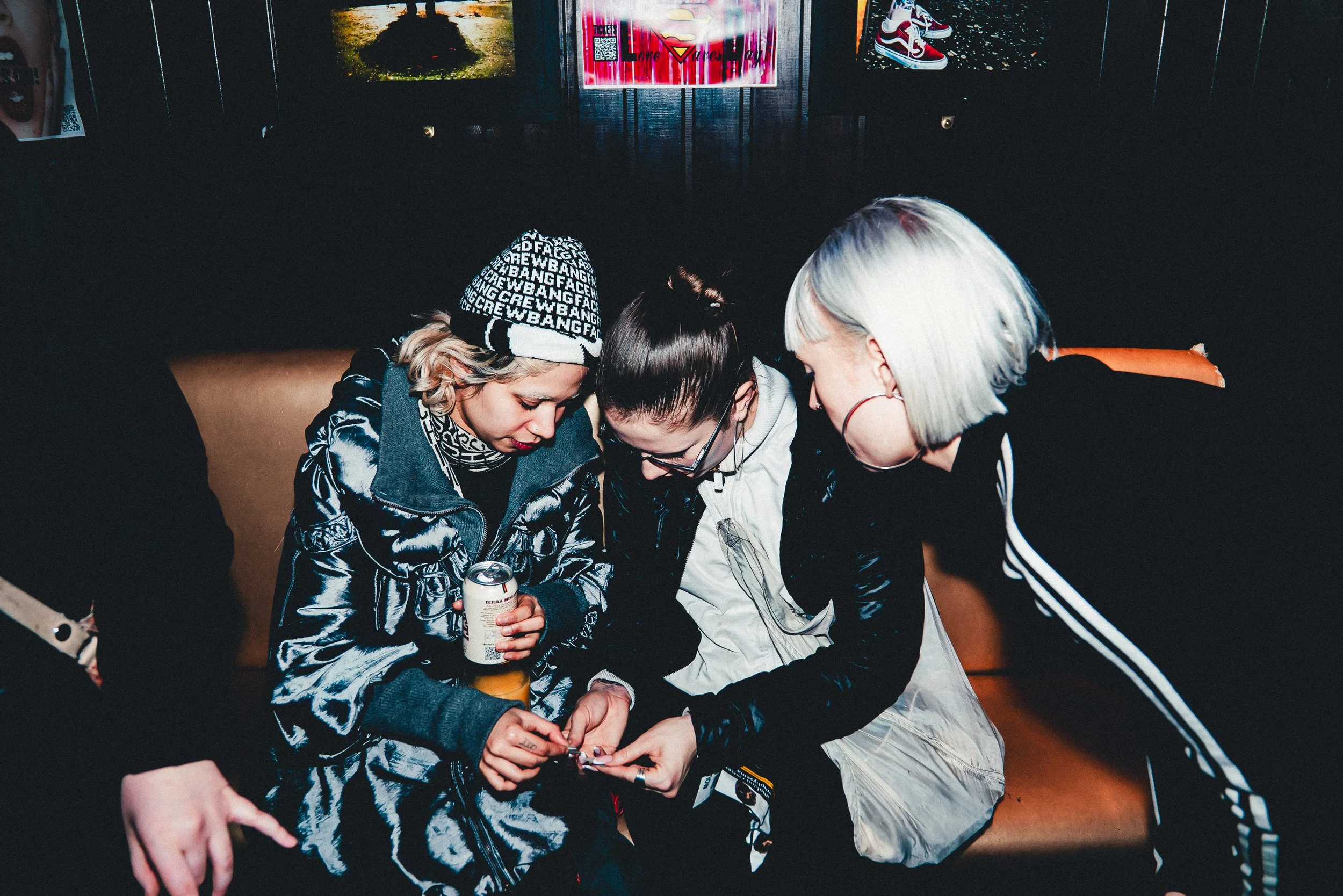 Three young women are sitting closely together on a couch, huddled around a smartphone. They are in a dark room with posters on the wall behind them, including a pair of sneakers and a colorful graphic. One woman is holding a can of soda, and all are