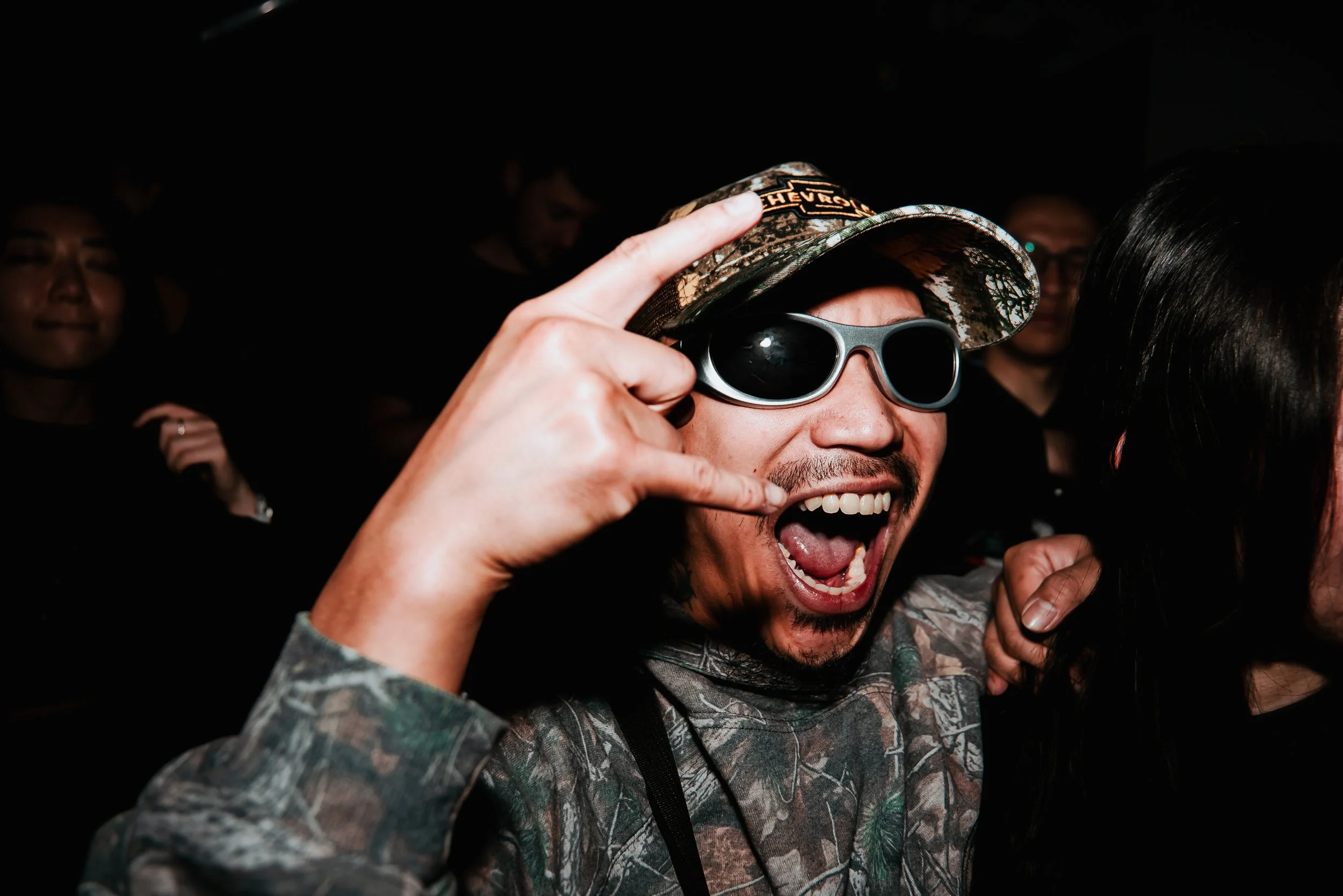 Man wearing camouflage hat and sunglasses making a rock and roll hand gesture and smiling widely at a dark event.