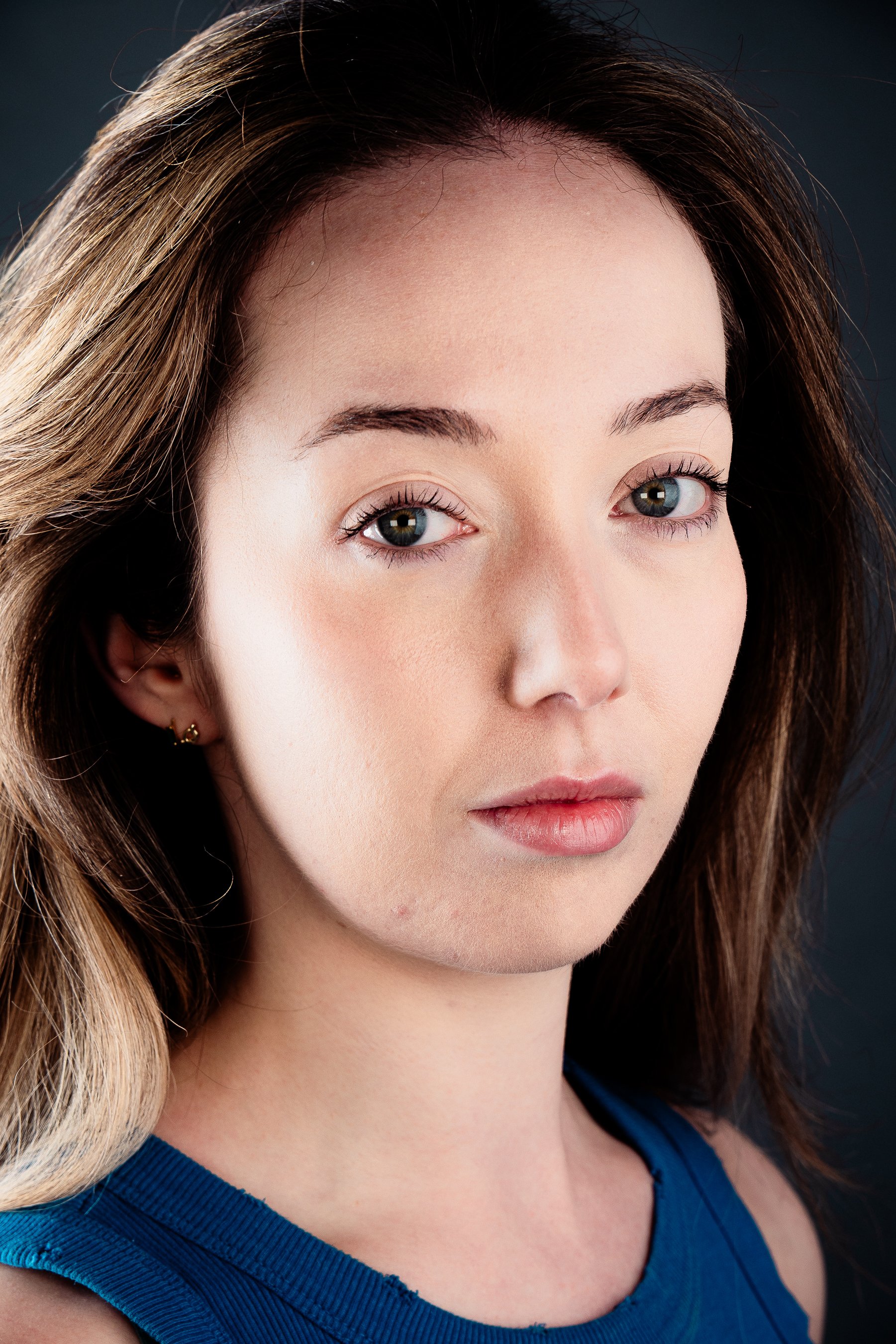 Close-up of a young woman with brown hair, blue eyes, wearing a blue top, and gold earrings, looking directly at the camera with a neutral expression.