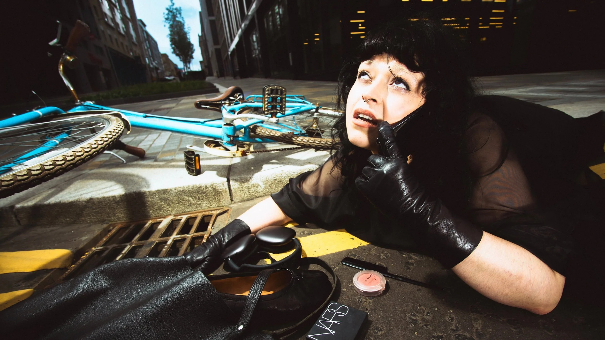A woman with black hair and a septum piercing lying on the ground near a bicycle, talking on a phone, with a black purse and makeup items nearby, in an urban setting at dusk.