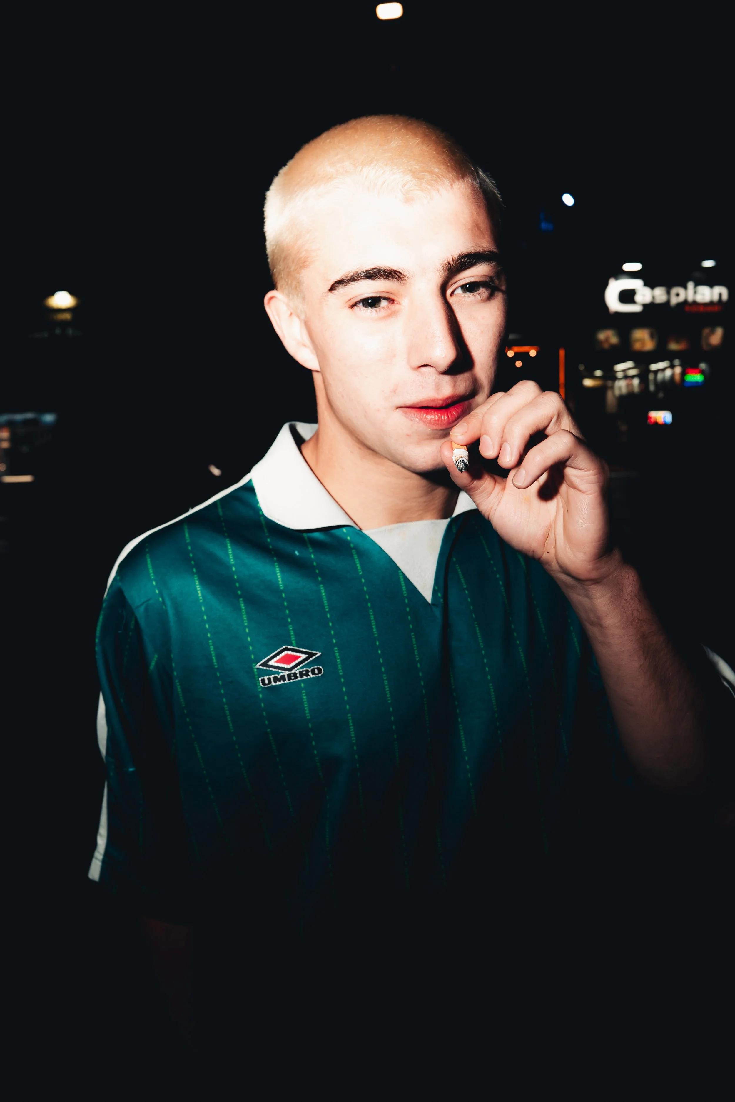 Young man with short blonde hair wearing a green and white jersey holding a cigarette in his hand, indoors at night with illuminated signs in the background.