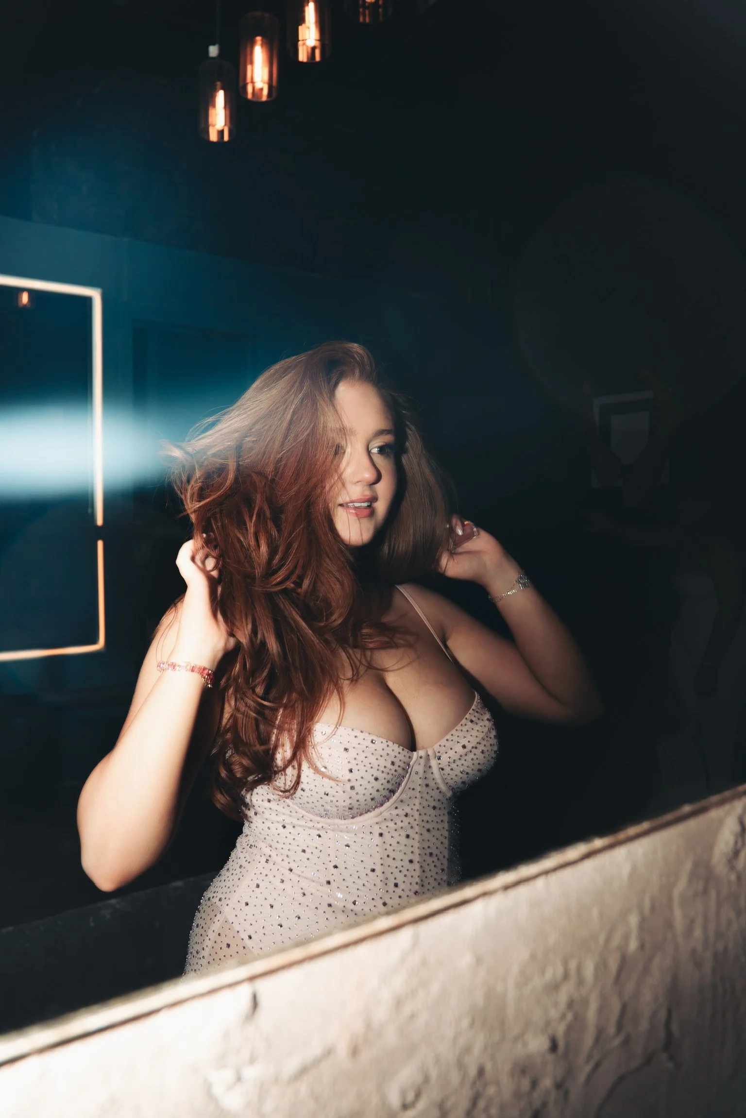A young woman with long, wavy red hair looks into a mirror while adjusting her hair in a dimly lit room, wearing a light-colored dress with small dark polka dots.