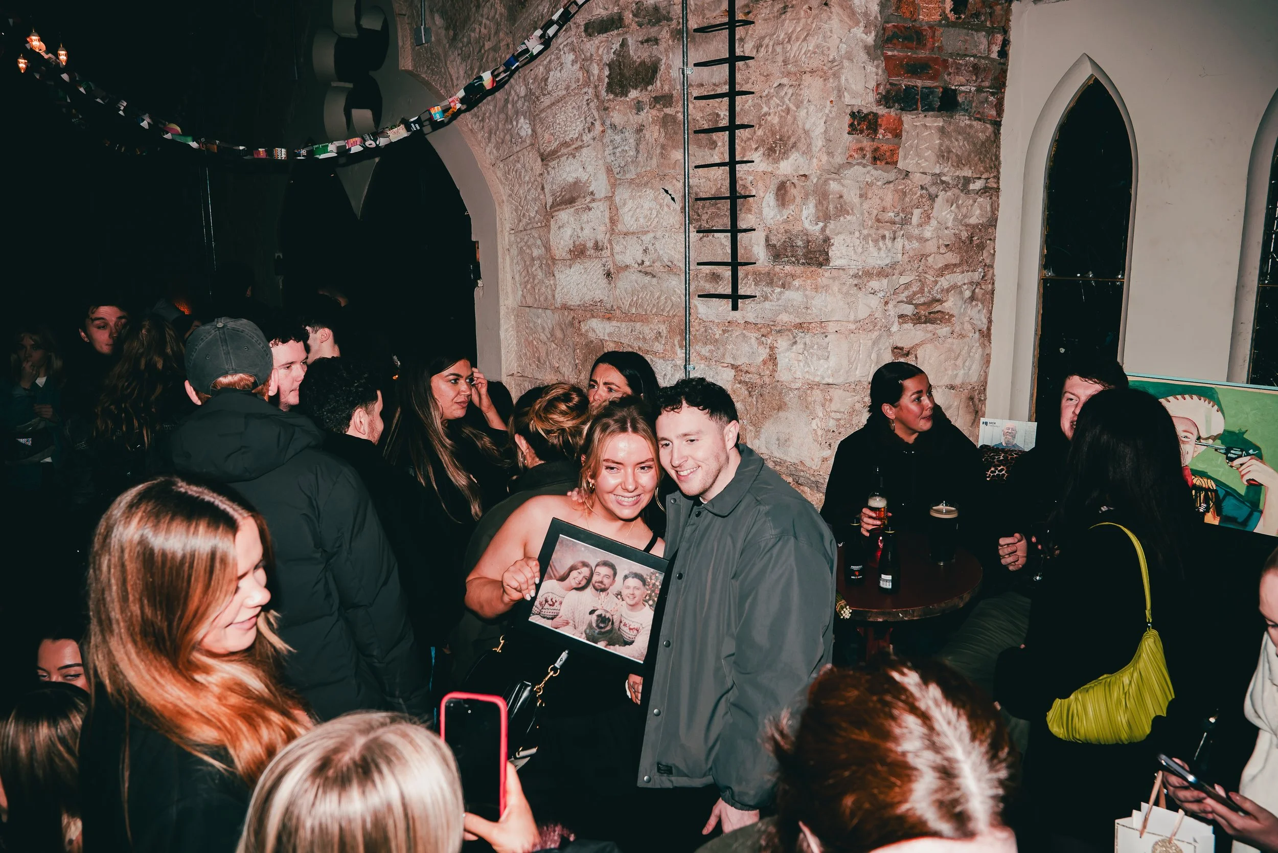 A group of people gathered closely together at a social event in a venue with exposed brick walls. Some are engaged in conversation while others are smiling and taking photos. One woman is holding a framed photograph of a group of friends. The scene 