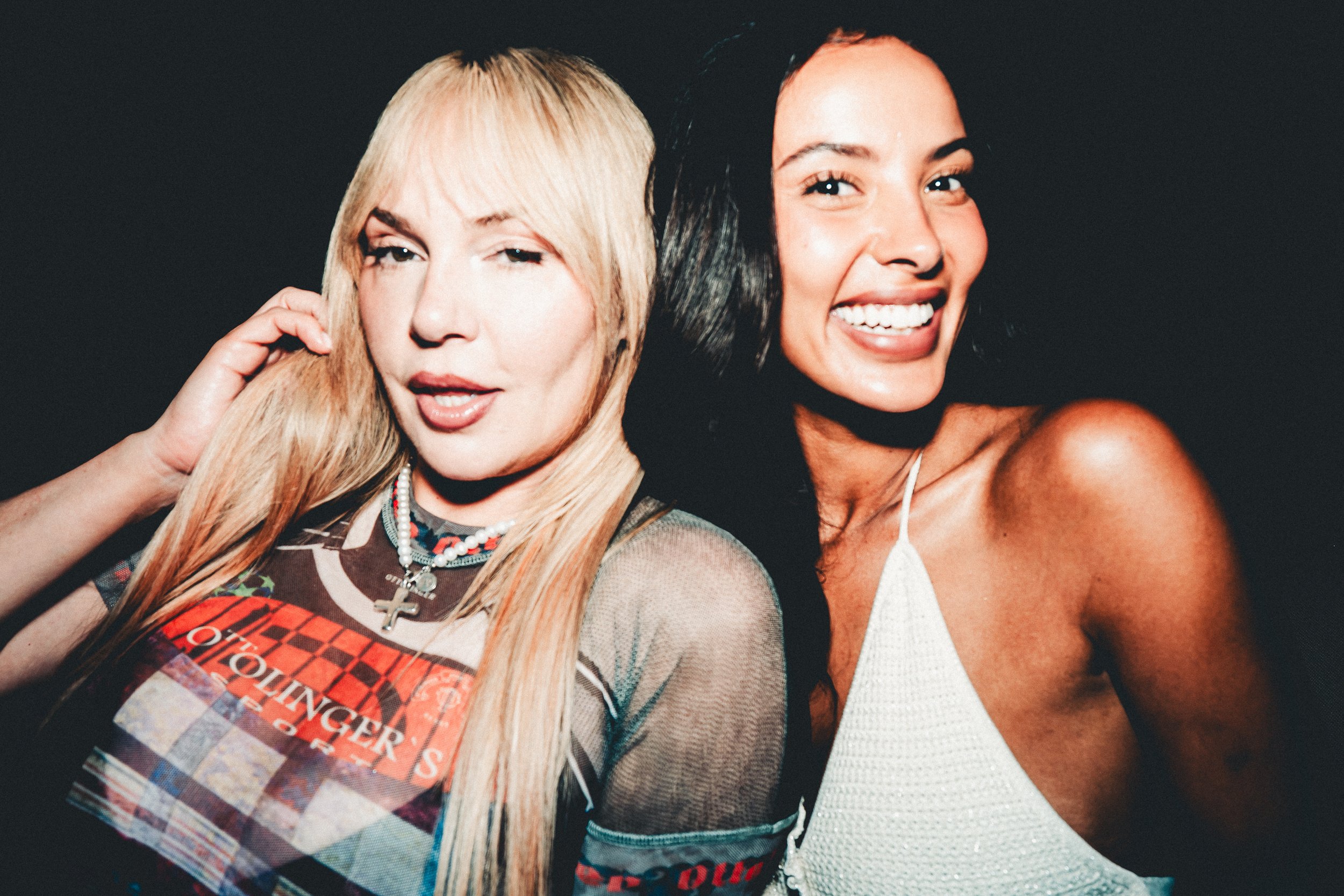 Two women smiling, close-up shot, with dark background. One has blonde hair and is winking, the other has dark hair and is showing teeth.