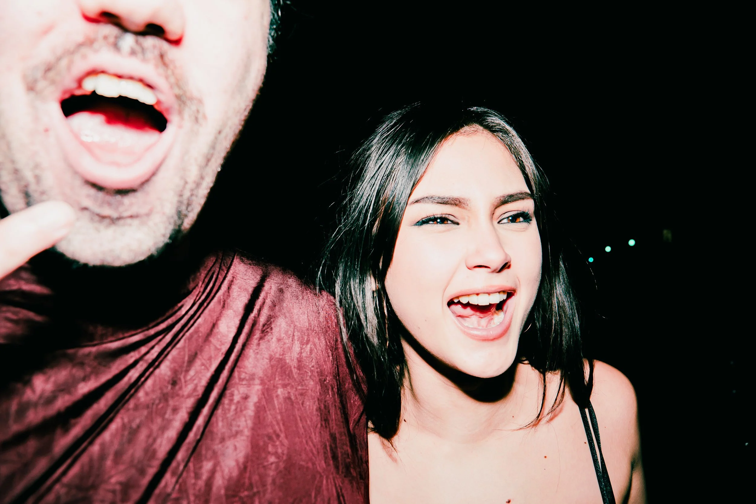 Close-up of a man and woman smiling and making silly faces with their mouths open, against a black background.