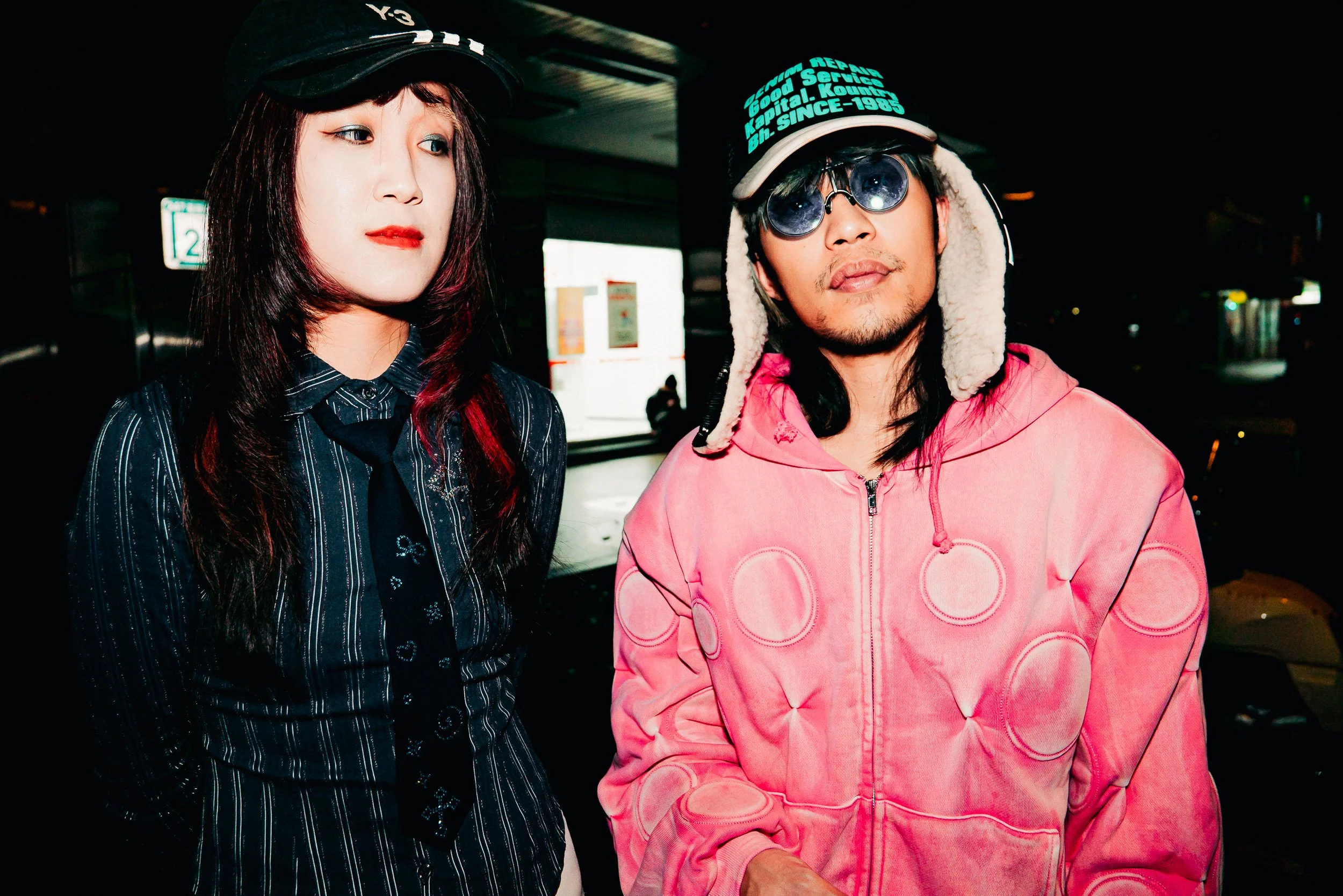 Two people, a woman with shoulder-length dark hair and red lipstick wearing a black cap and a striped shirt, and a man with shoulder-length dark hair, sunglasses, a black cap with text, and a pink hoodie, standing together in a dimly lit indoor space