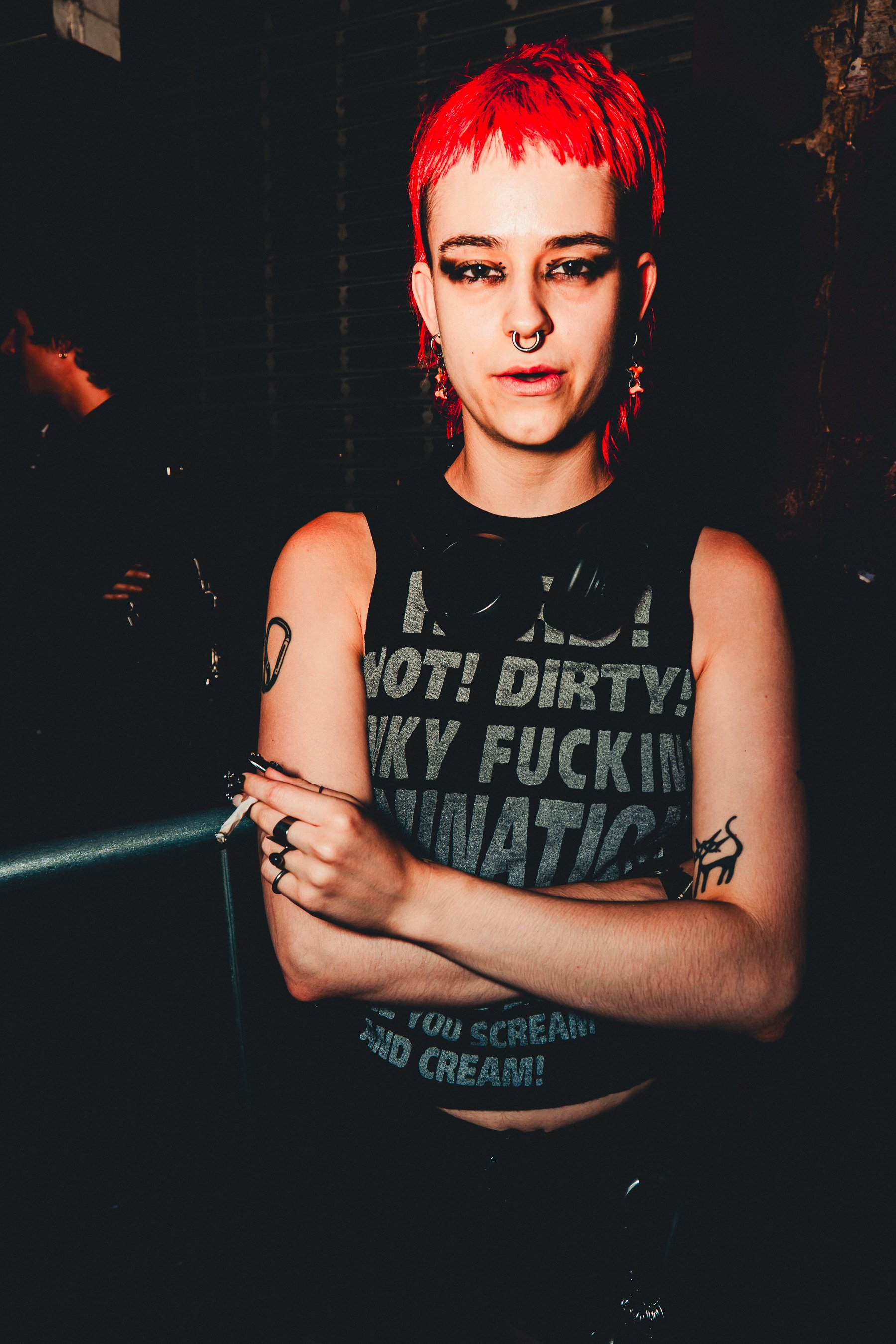 A person with short red hair, multiple facial piercings, and tattoos is standing with their arms crossed, wearing a sleeveless shirt with bold black and gray text. The background is dark, with some indistinct figures and structures visible.