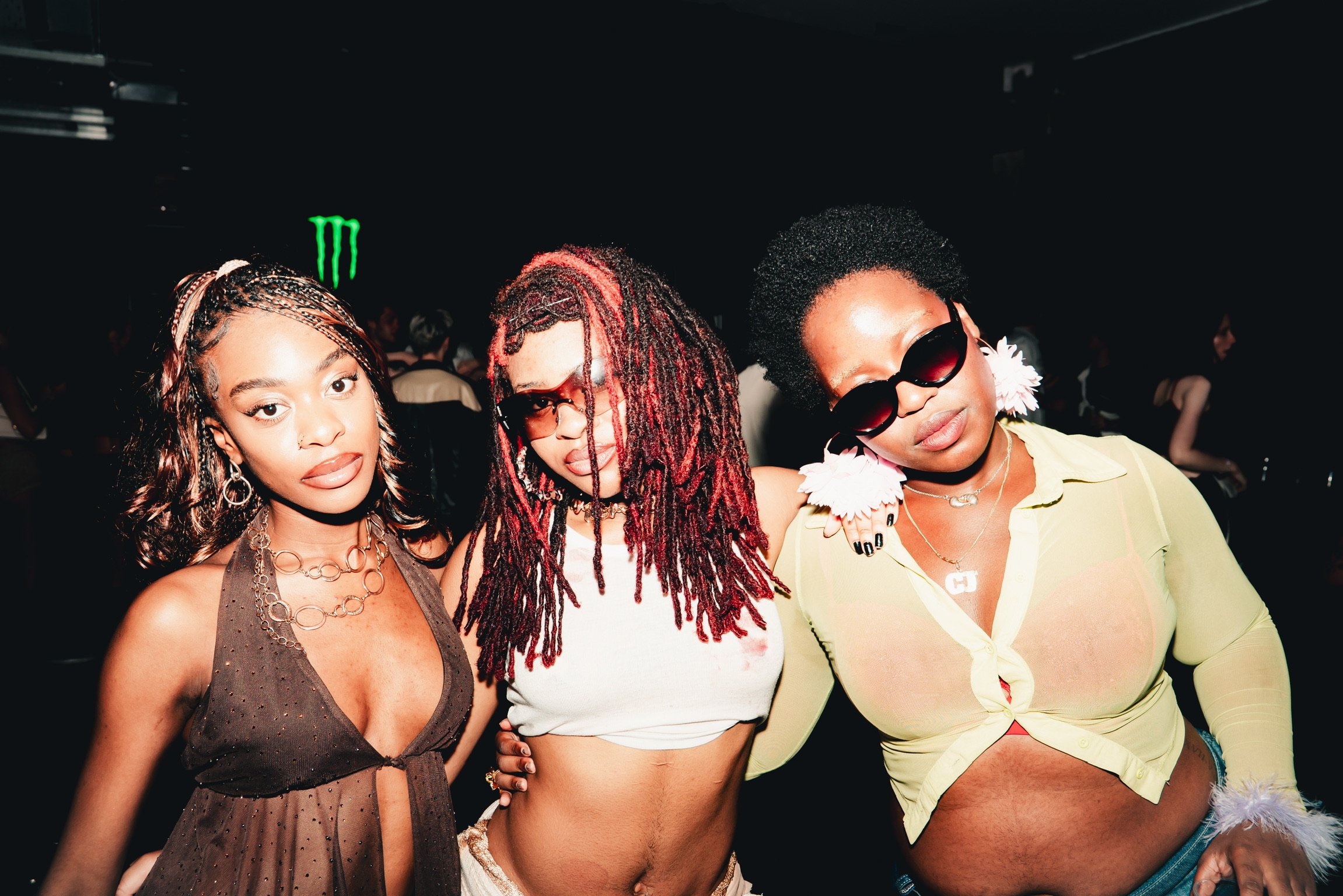 Three women posing together at a social event, with dark background and a neon green monster energy logo visible.