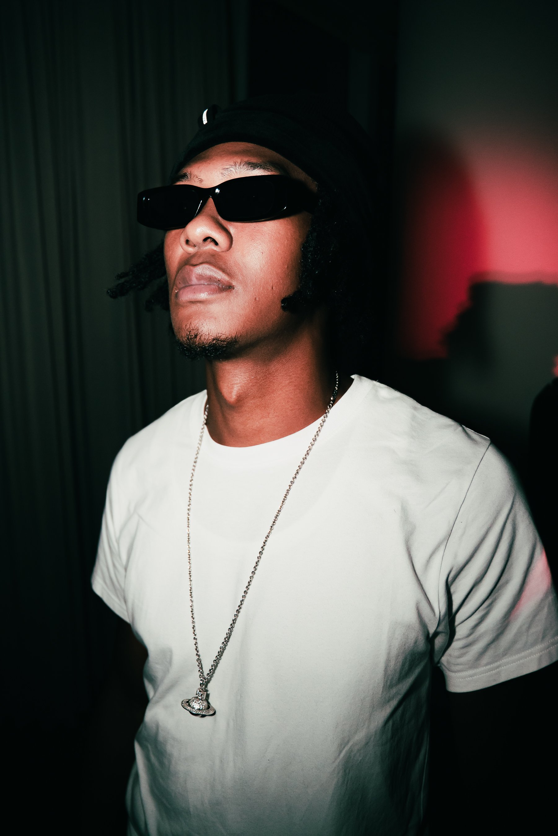 A person wearing black sunglasses, a black cap, a white T-shirt, and a silver chain with a pendant, standing in a dimly lit environment with a dark background and a red light source.