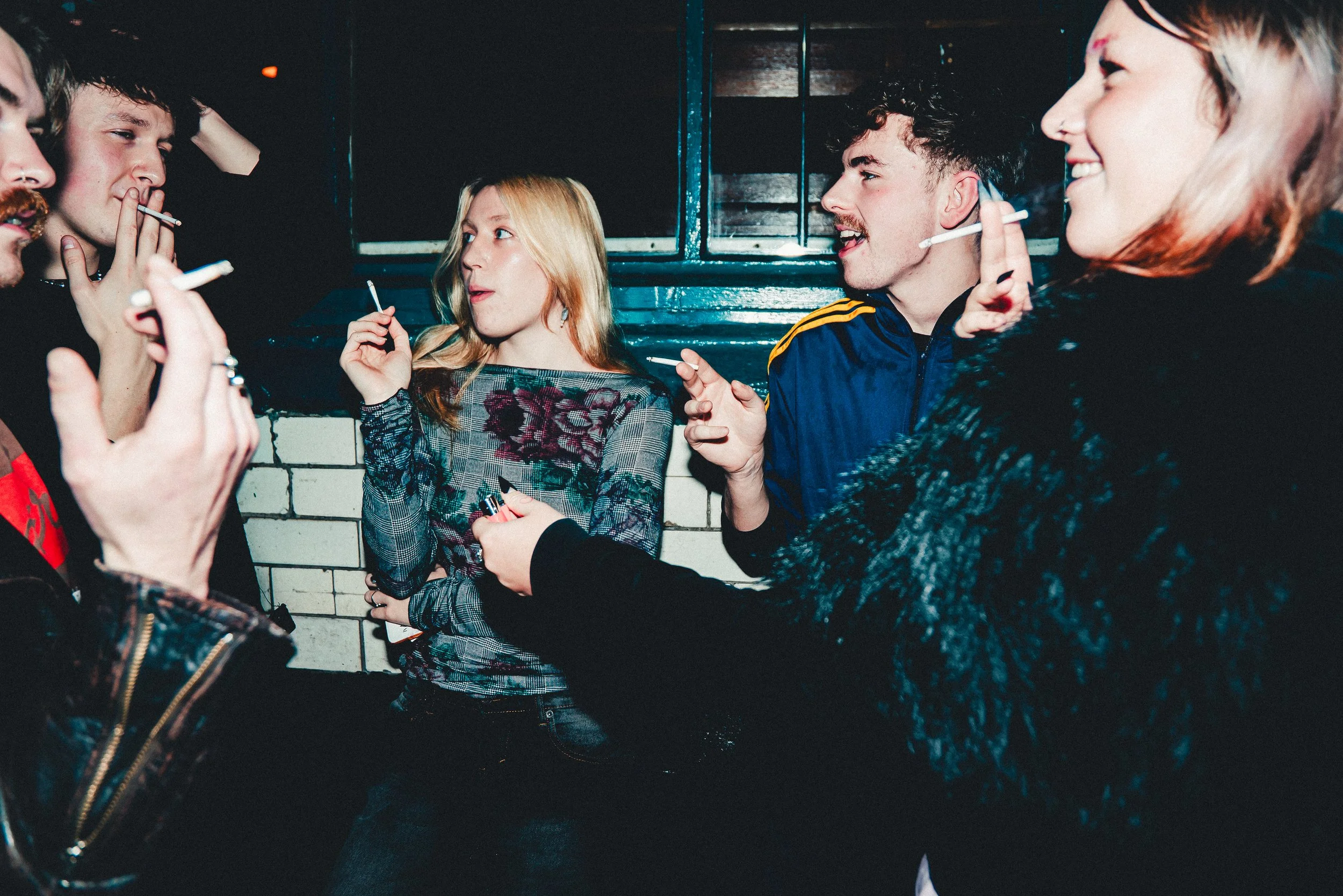 Group of young people socializing indoors, smoking cigarettes, chatting and smiling.