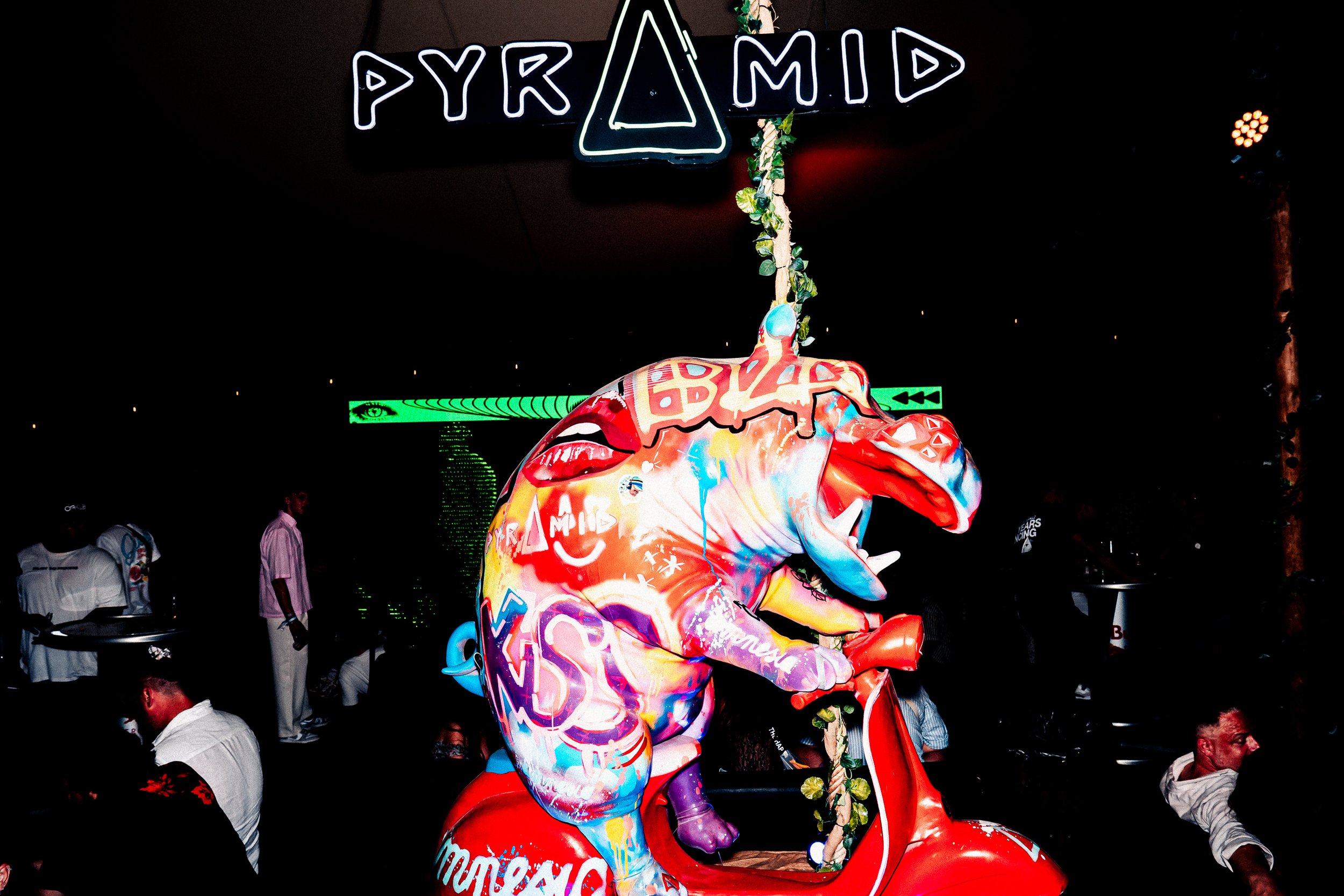 Colorful mannequin of an elephant with graffiti-style art, surrounded by people at a nighttime event, with a neon sign reading 'PYRAMID' above.