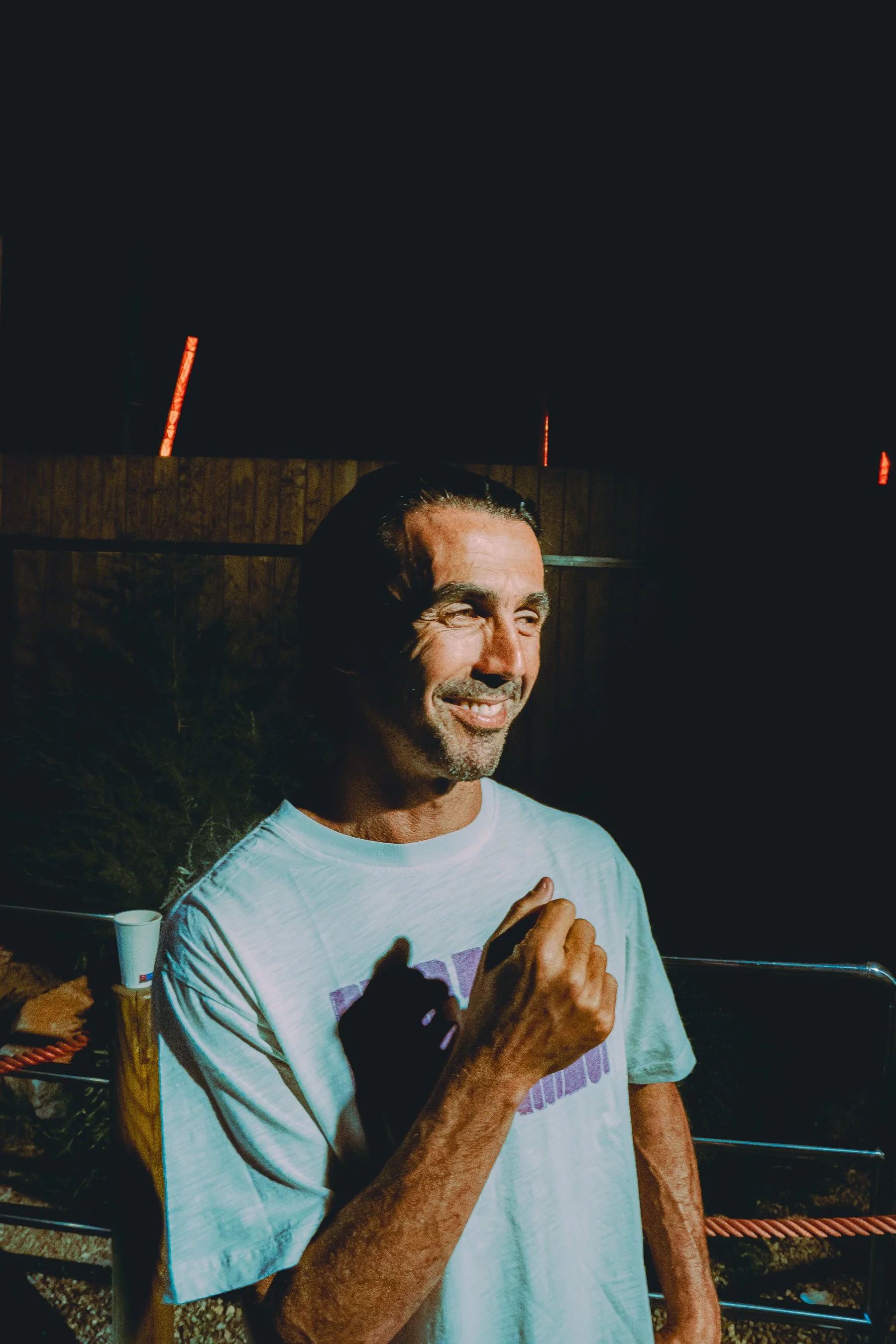 A man with dark hair and a goatee smiling outdoors at night. He's wearing a light-colored T-shirt and holding his right hand near his chest. Behind him is a wooden fence and some red lights.