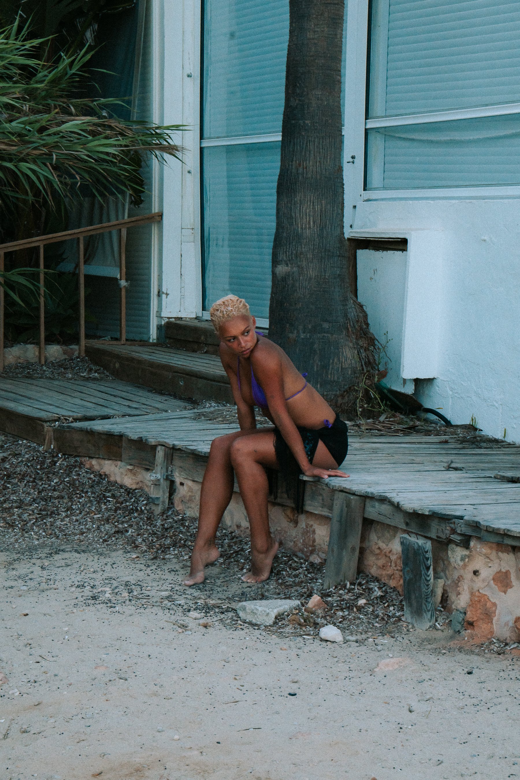 A woman with blonde hair sitting on a wooden platform outside near a tree, wearing a purple top and black shorts, with a white building and window with closed blinds in the background.