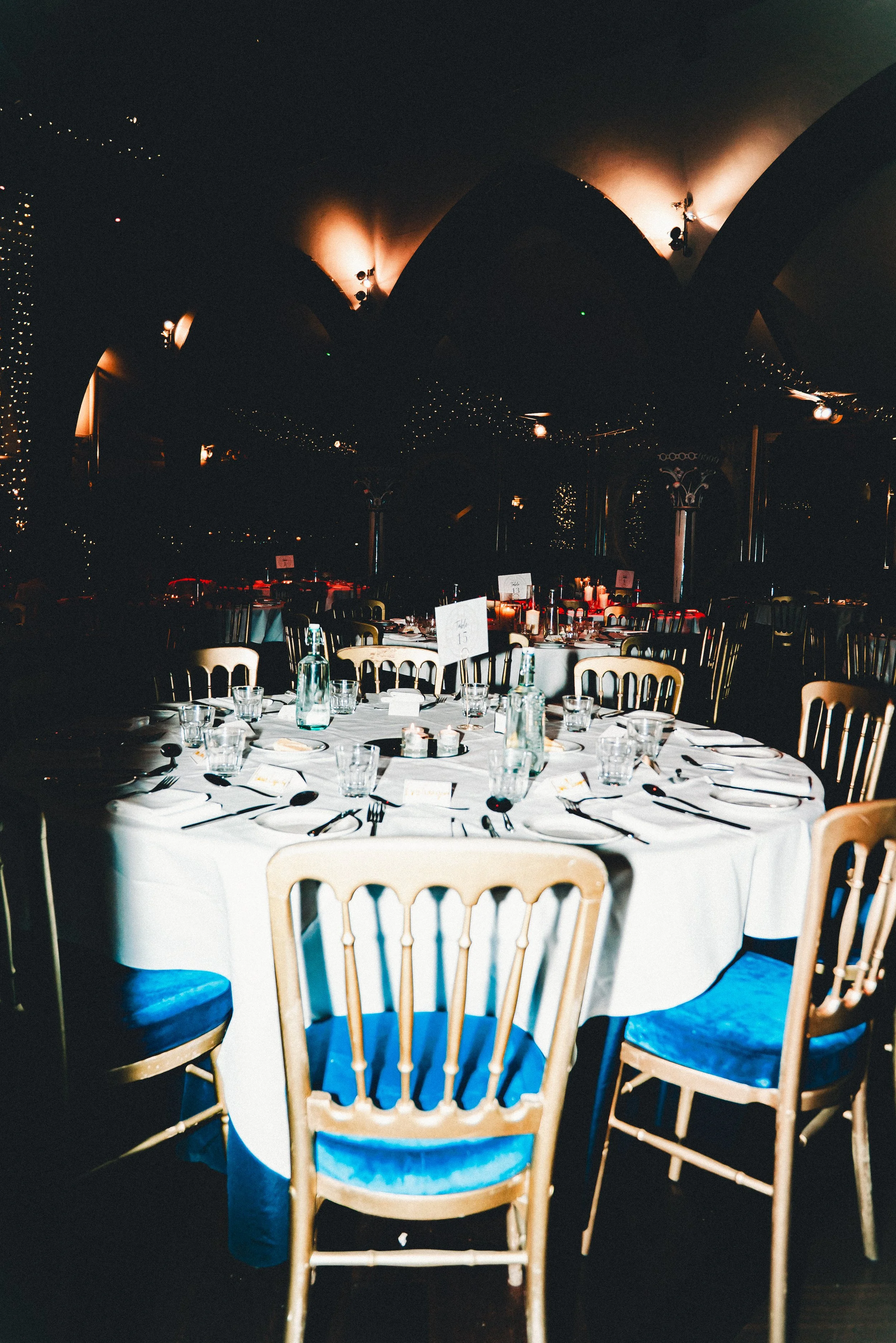 A large round dining table set for a formal event with white tablecloths, glass bottles, and place settings. Wooden chairs with blue cushions surround the table. In the background, there is a dark ceiling with warm lighting and string lights, creatin