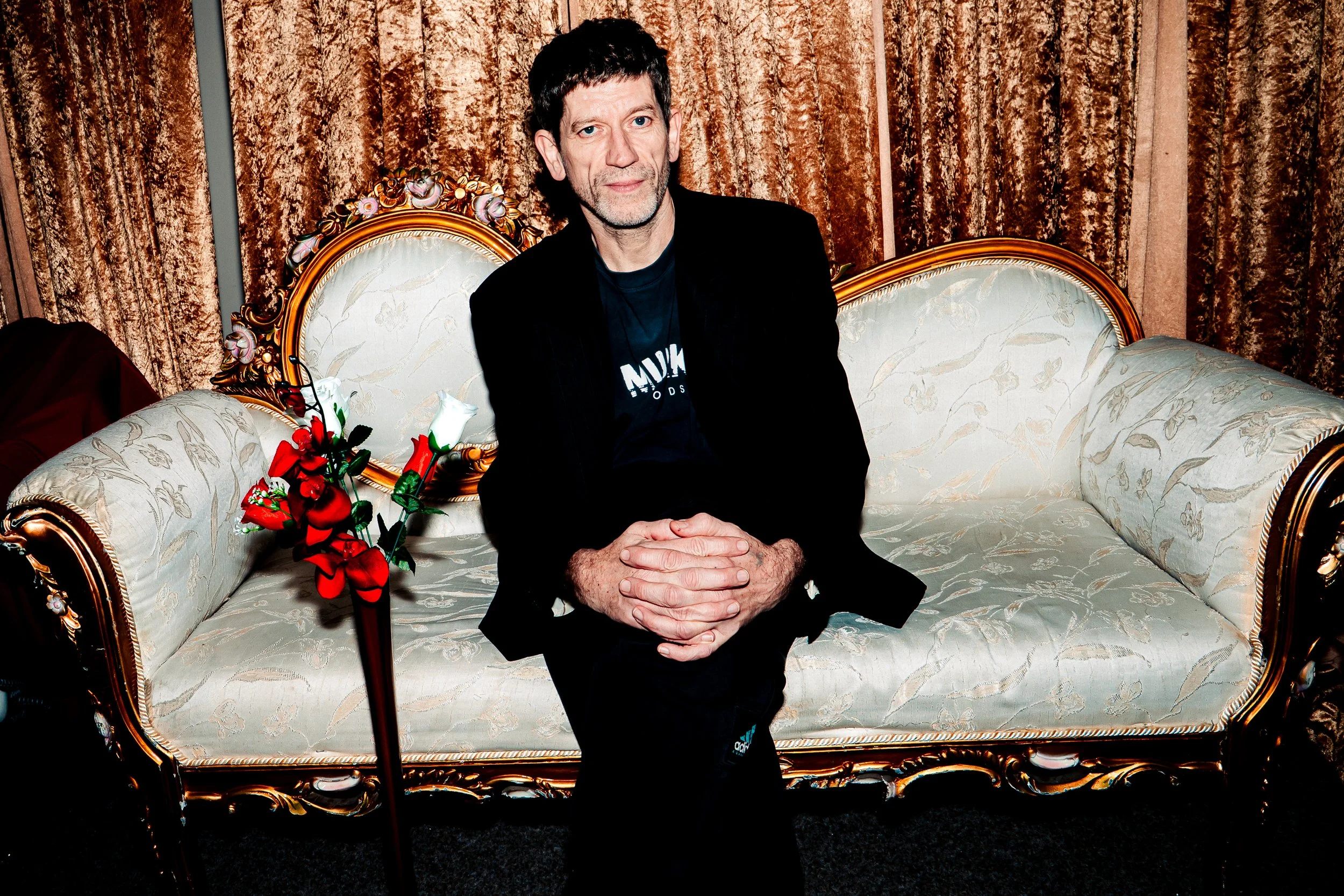 A man with dark hair and a beard sitting on an ornate cream-colored vintage sofa with gold accents, in front of gold-brown curtains, holding his hands together with a festive bouquet of red and white flowers on his side.