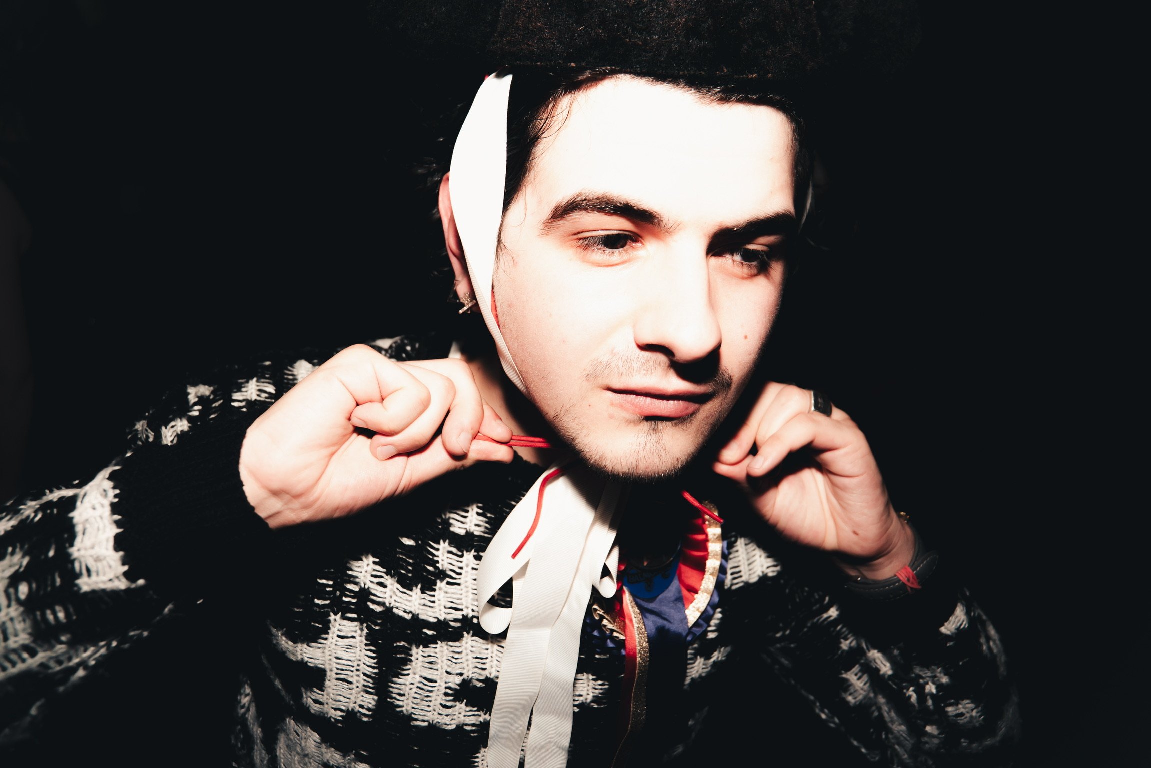 A young man with dark hair and earrings wears headphones and a patterned sweater, adjusting the headphones around his neck.
