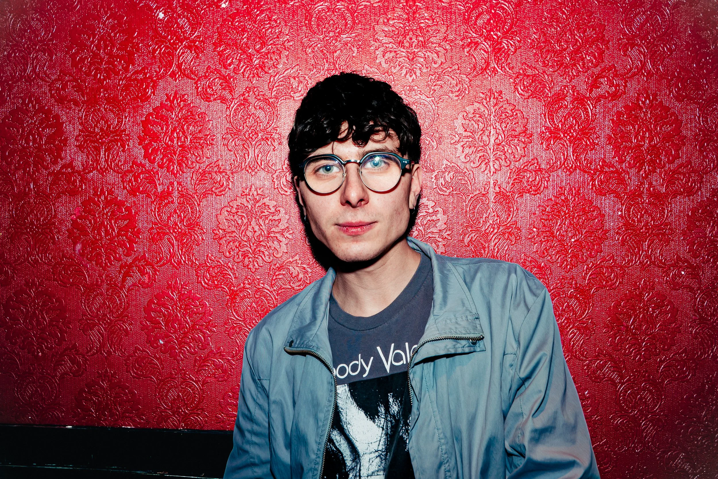 A young man with curly black hair, wearing glasses and a blue jacket, standing in front of a red textured wallpaper background.