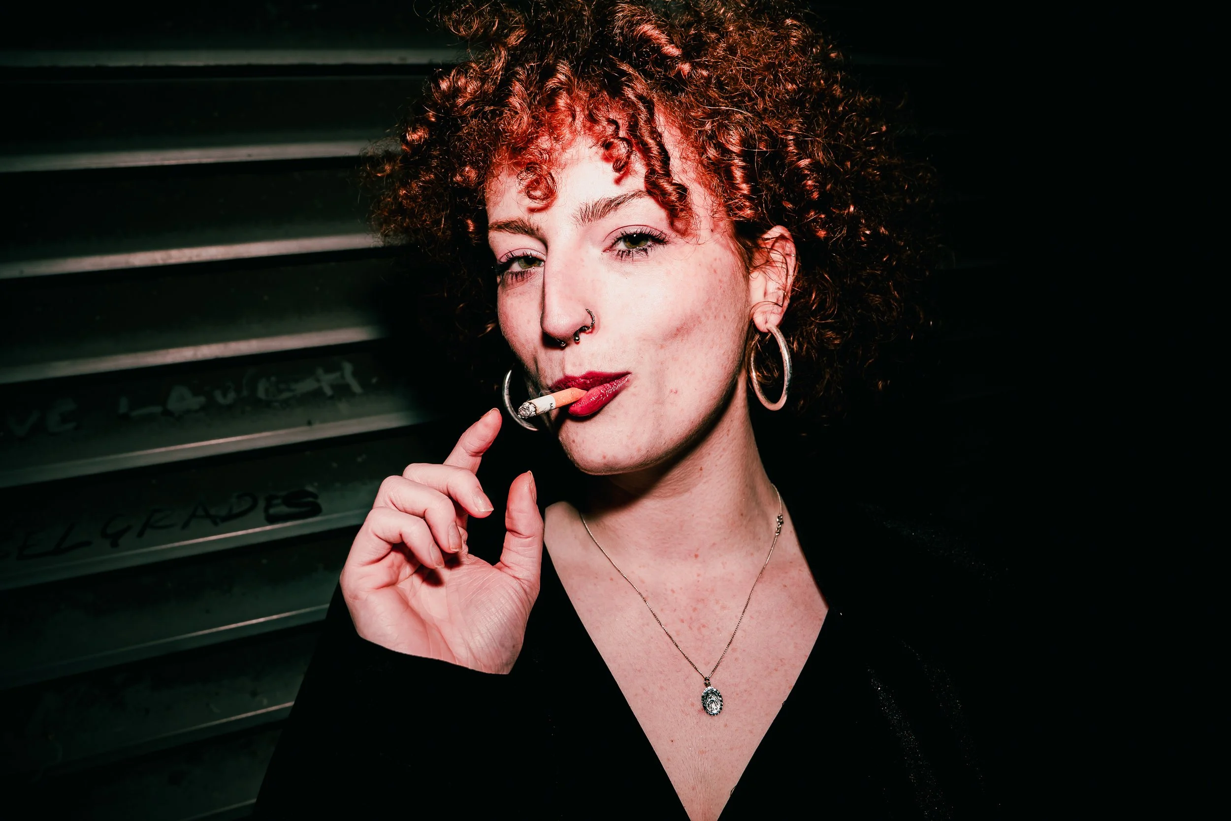 A woman with curly red hair and fair skin is holding a cigarette to her lips while looking at the camera. She is wearing hoop earrings, a pendant necklace, and a black top. The background appears to be a dark room with some metallic objects and handw