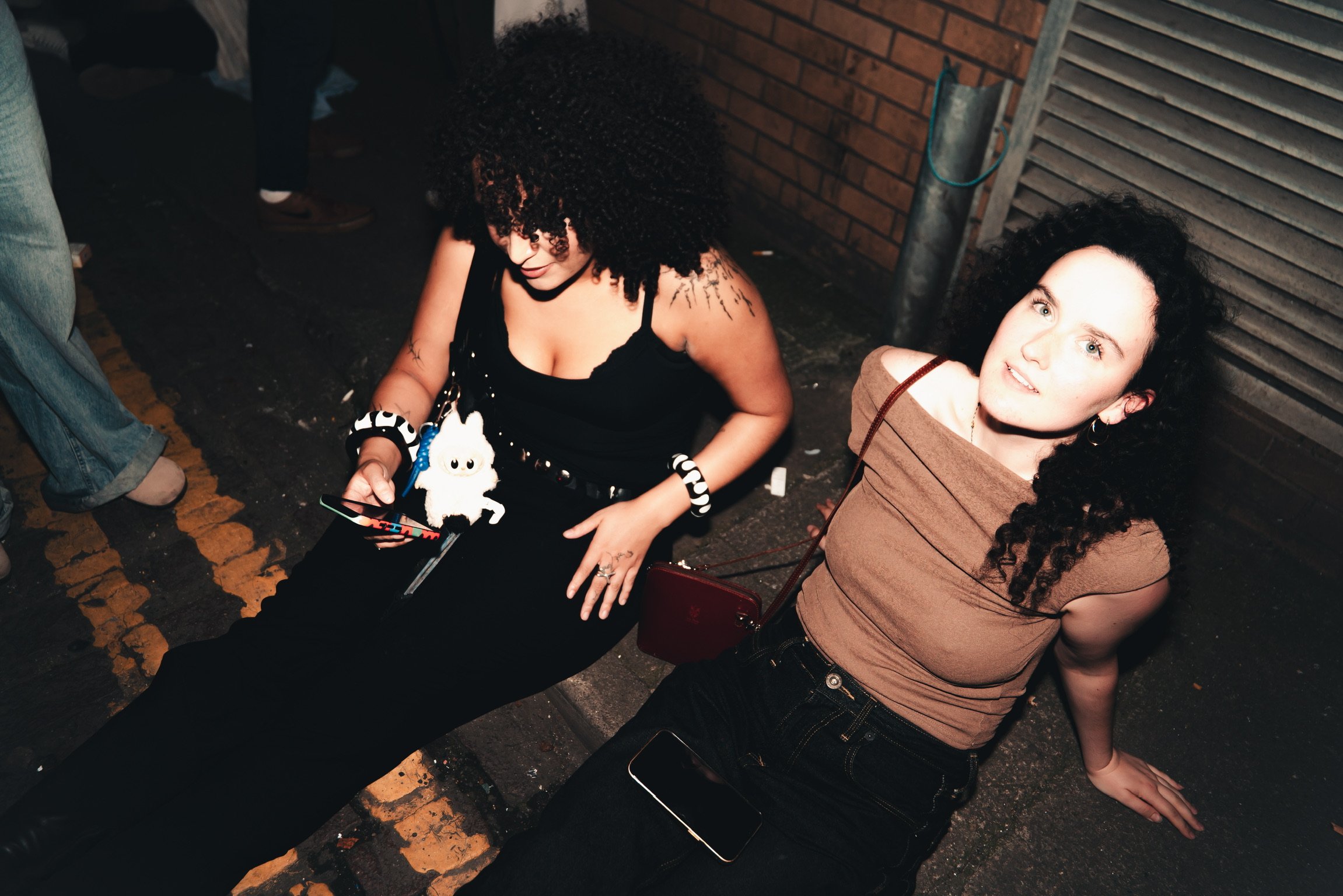 Two women with curly dark hair sitting on the ground against a brick wall at night, one looking at her phone, the other looking upward with a neutral expression.