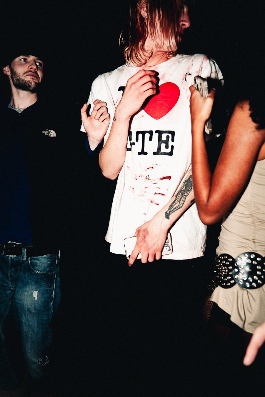 Person with dyed hair wearing a torn white T-shirt with a red heart and black text, holding a phone, standing next to a woman with a tattooed arm and studded belt in a dark environment.