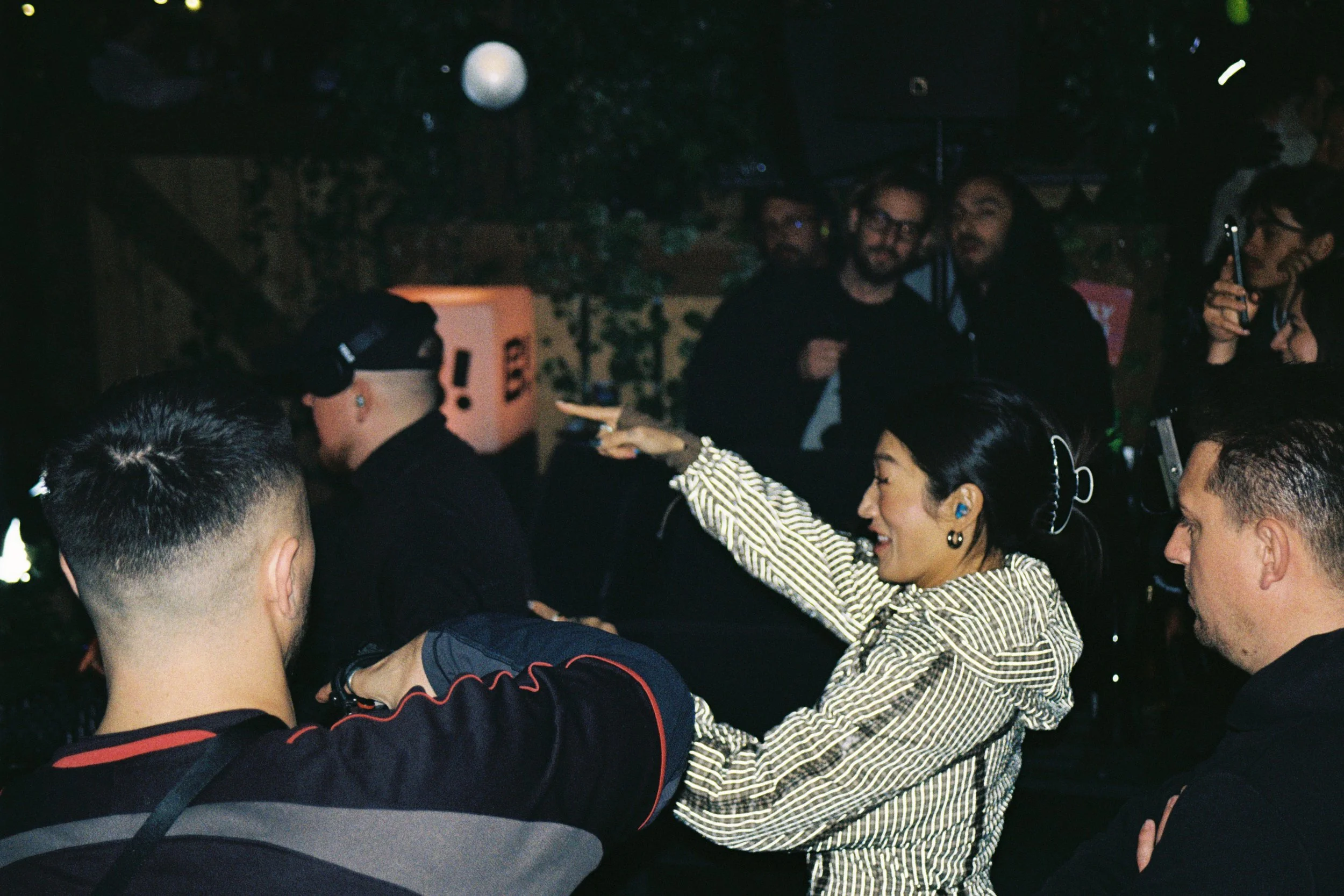 A group of people gathering indoors, some wearing headphones, with a woman smiling and reaching out to a man, and others engaging with electronic devices.
