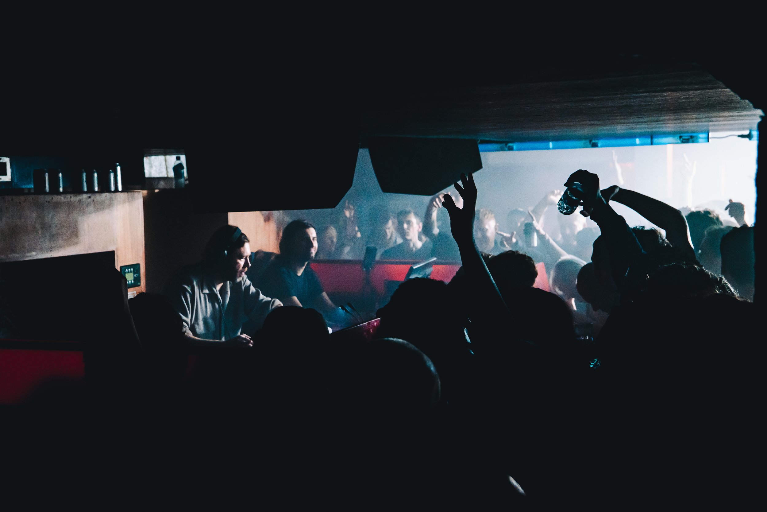 People dancing and enjoying music in a dark nightclub with DJ and colorful lighting.