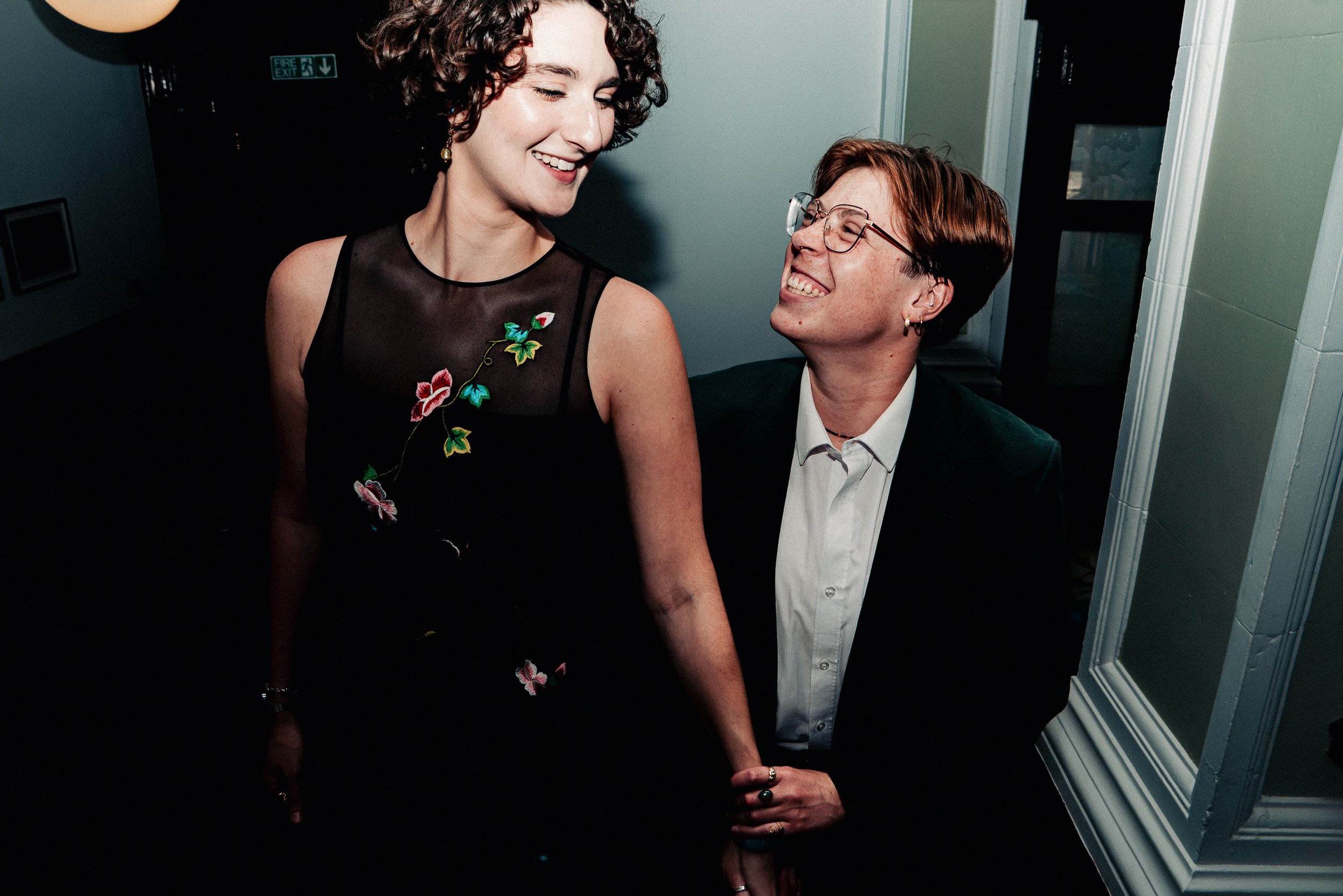 Two women smiling and looking at each other, standing close together indoors near a door with a window, one wearing a black sheer top with floral embroidery and earrings, the other with glasses, short hair, and multiple rings.