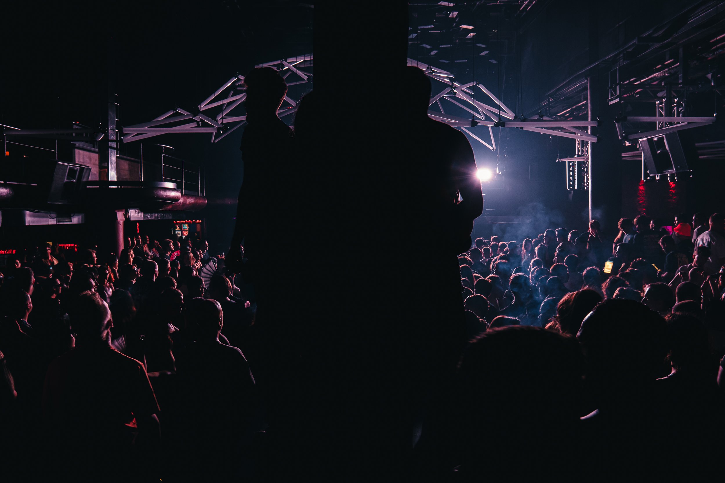 A crowded indoor event with silhouettes of people in the foreground, and a stage with bright lights and geometric structures in the background.
