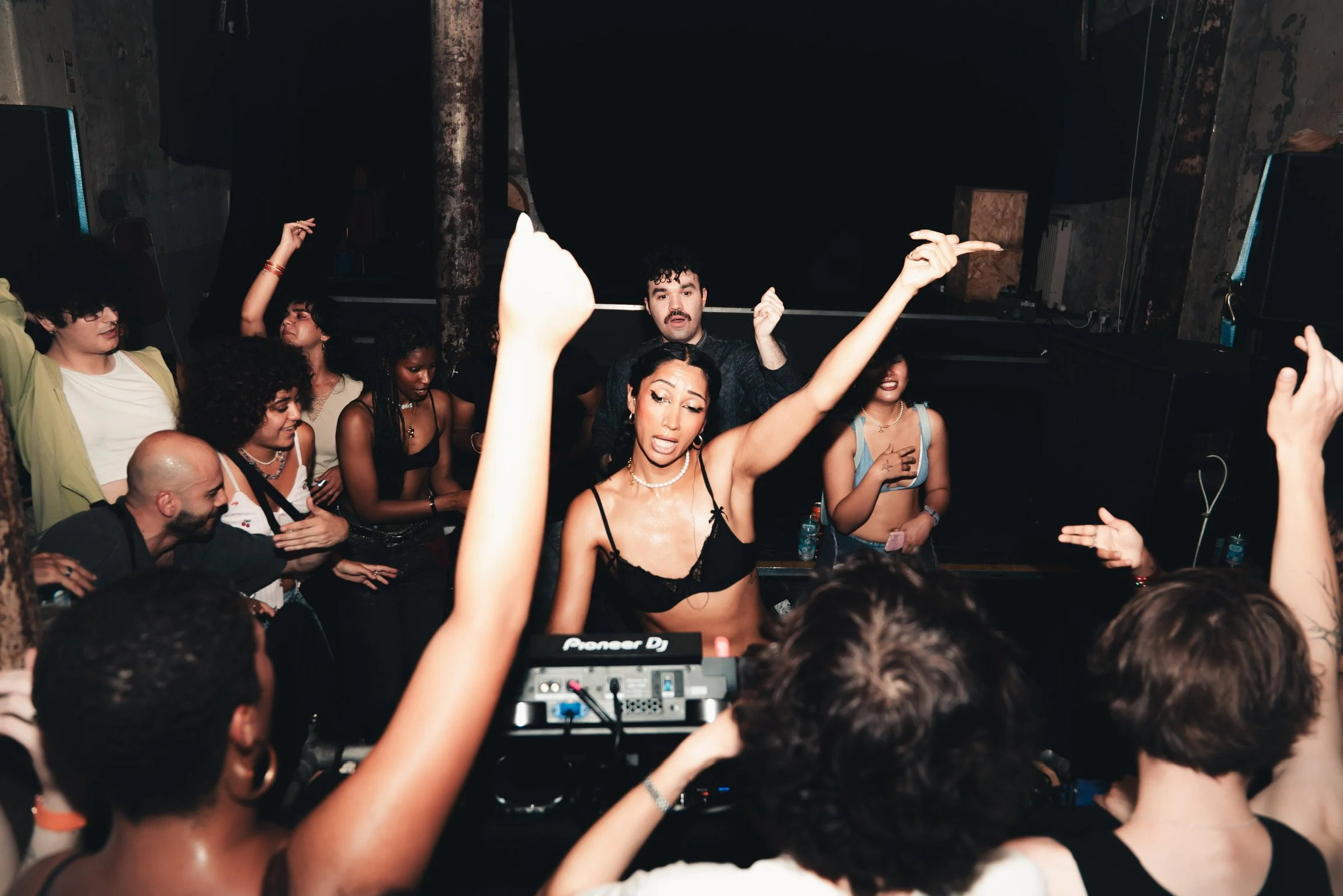 A crowded nightclub scene with people dancing and enjoying music, a DJ setup in the foreground, and a woman with long dark hair and a black top dancing with her arms raised.