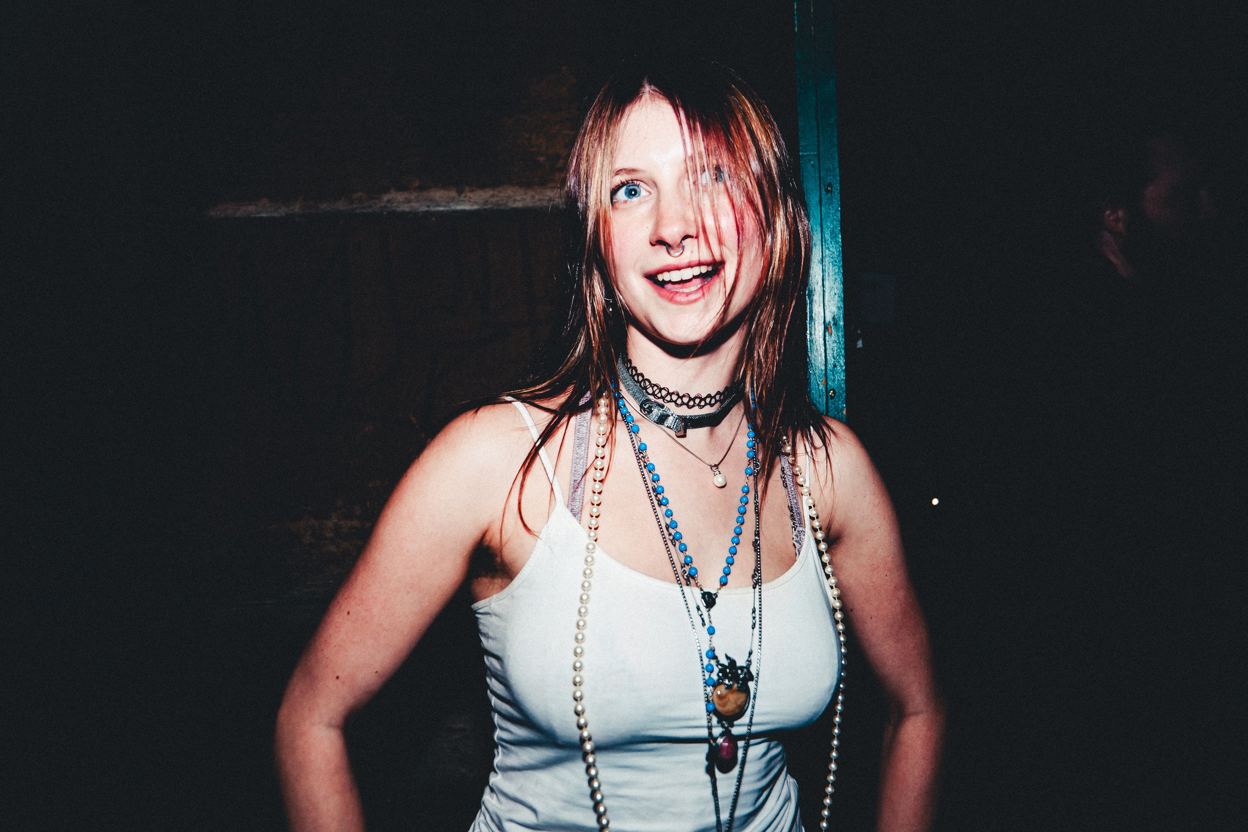 Young woman with red hair and blue eyes smiling and showing a septum piercing, wearing multiple necklaces, a white tank top, and standing against a dark background.