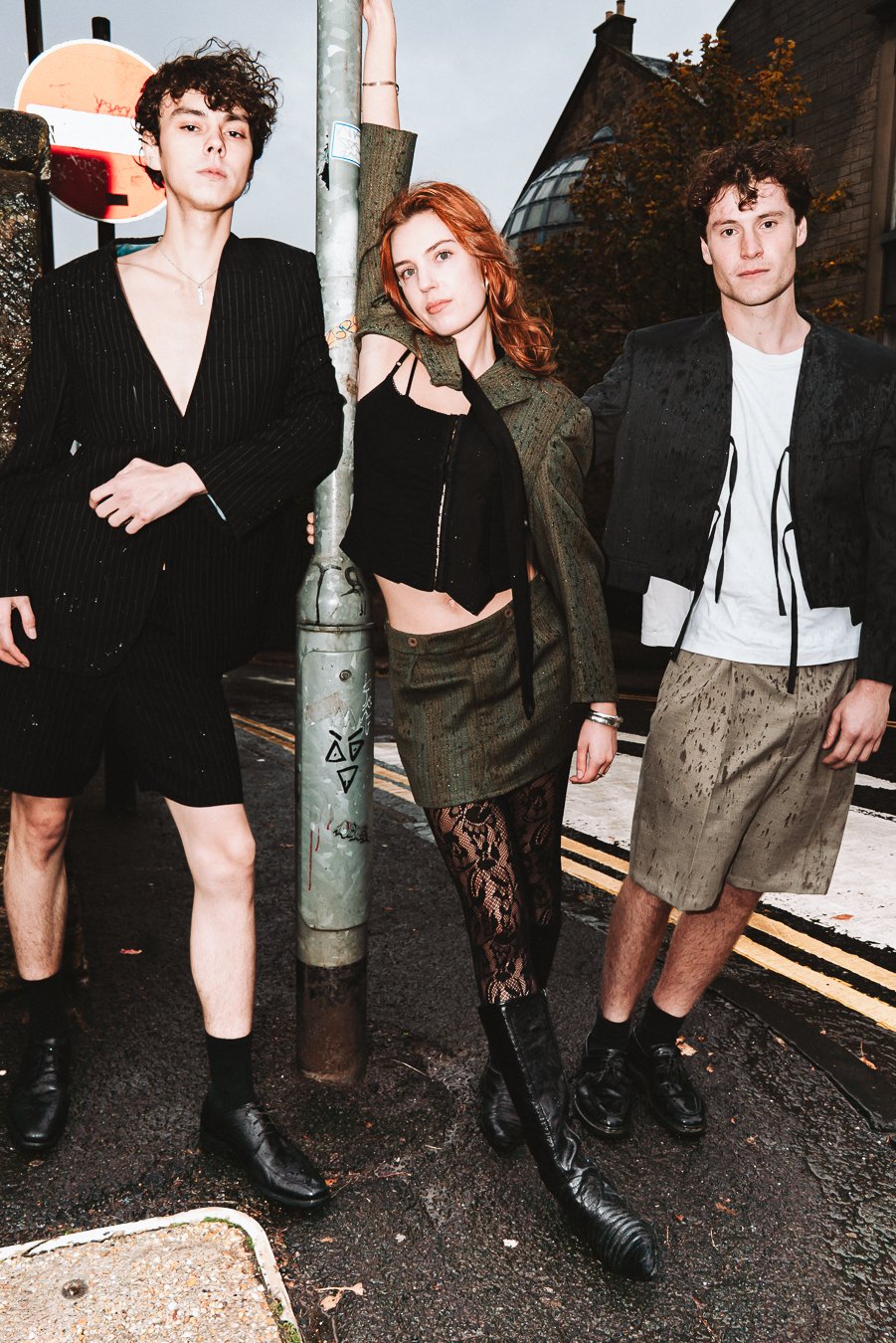 Three young individuals standing on a city street, leaning against a pole, dressed in fashionable, casual clothing, with an urban background, and one of them is wearing patterned tights.