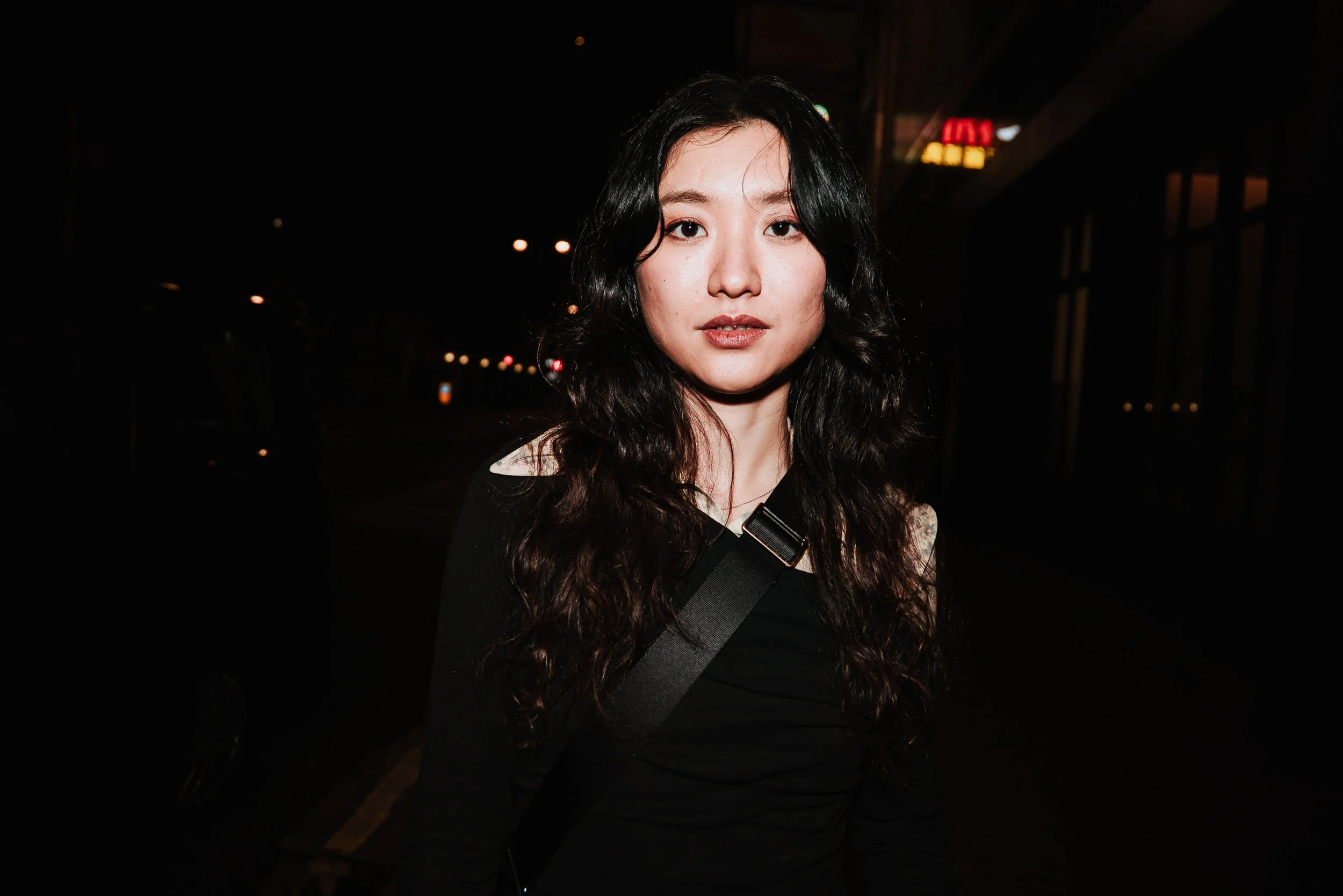 A woman with long black hair and light makeup standing outside at night, wearing a black top and a strap across her shoulder.
