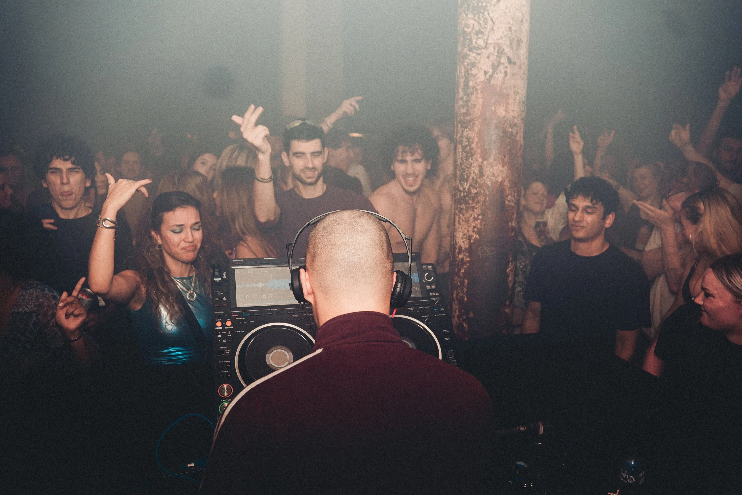 Crowd of people dancing and enjoying music at a concert or nightclub, with a DJ playing in the foreground.