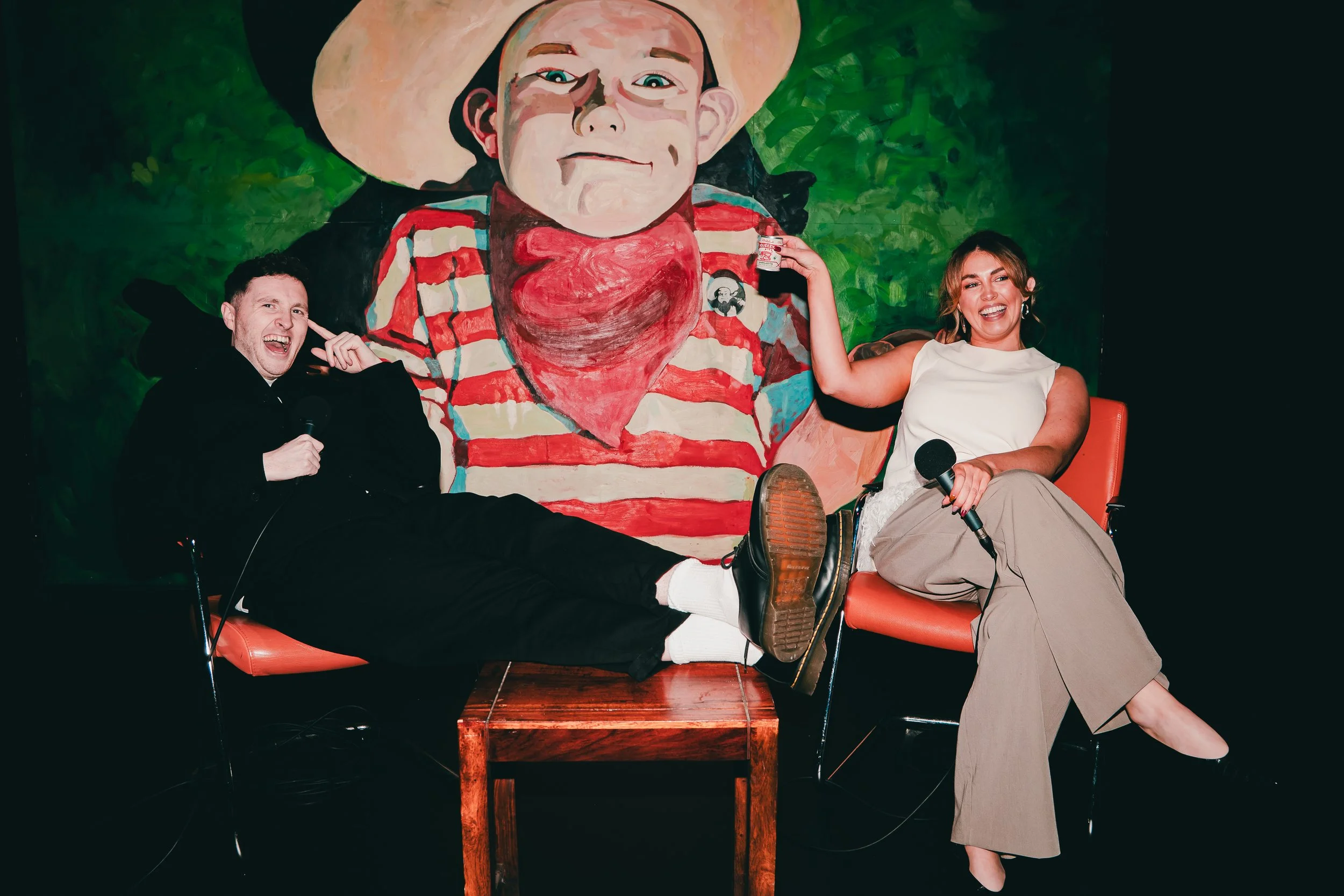 Two people, a man and a woman, sitting on orange chairs with a colorful mural of a cartoonish boy in a striped shirt and cowboy hat behind them. The man is wearing black and is holding a microphone, while the woman is wearing a white top and beige pa