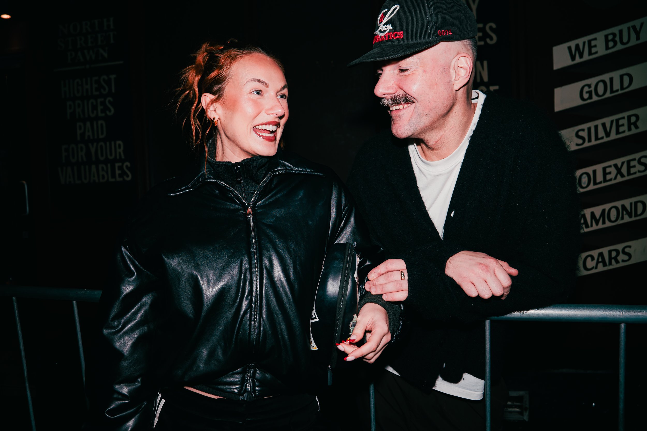A woman with red hair and a man with a mustache smiling and engaging in conversation at night, standing near a black fence with signs in the background.