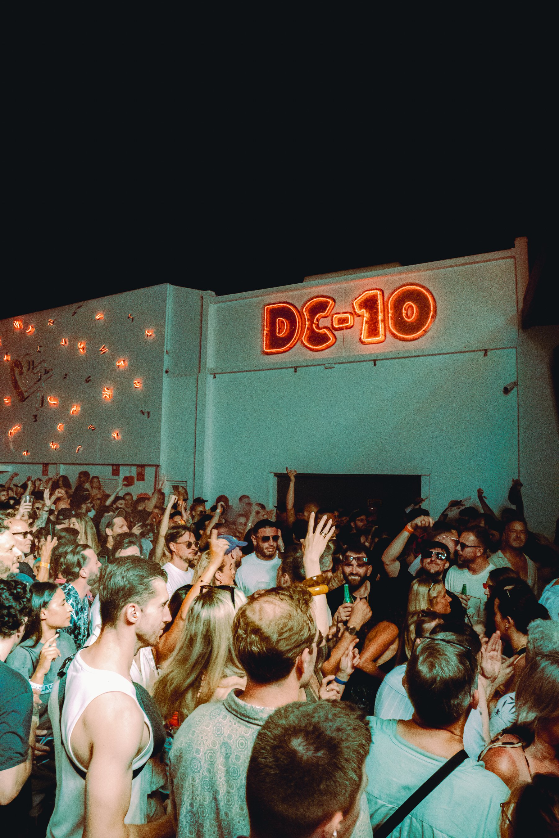 Crowd of people at an outdoor concert at night with a neon sign that reads 'DG-10' on a building behind them.