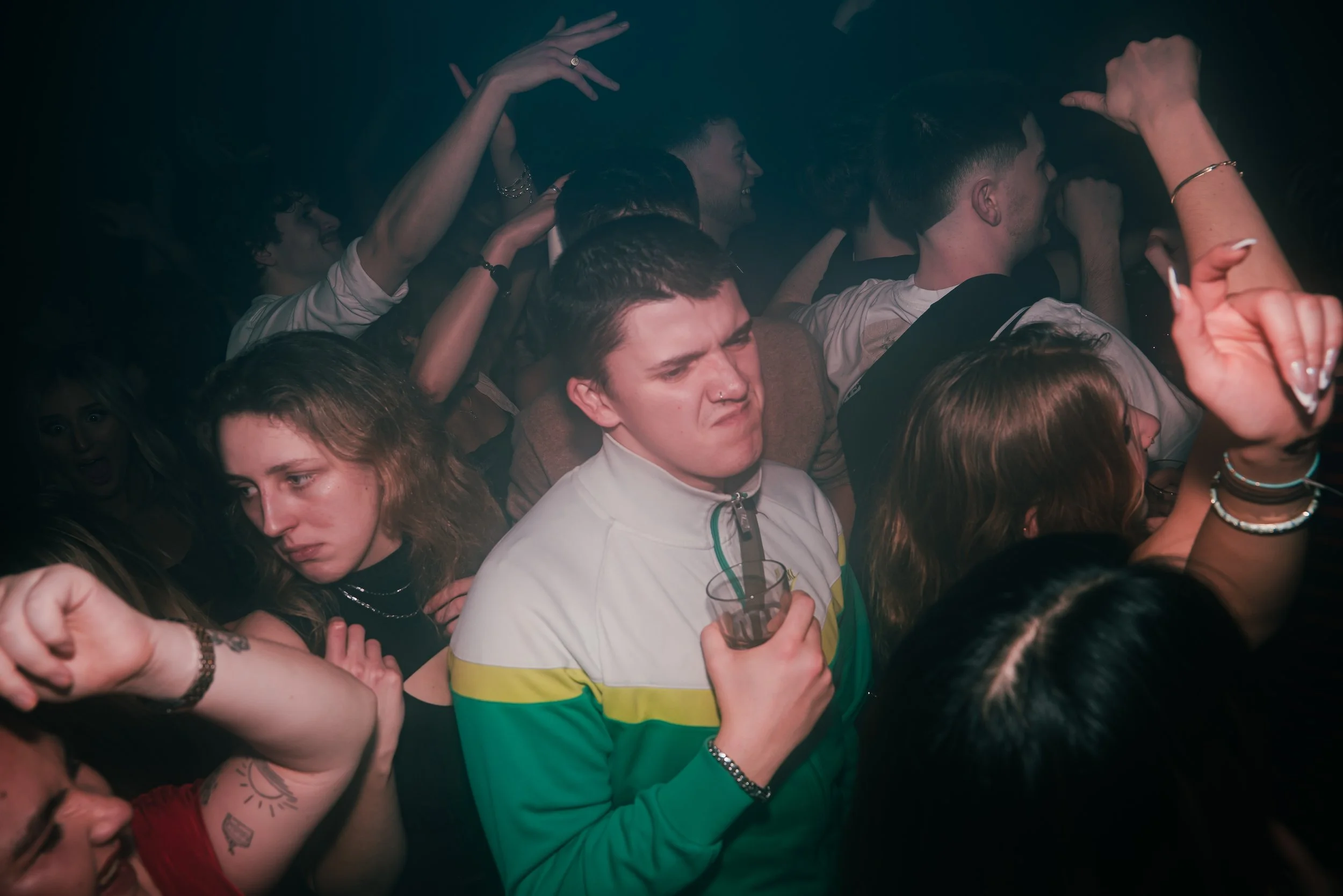 Crowd of people at a party or concert, some dancing and others holding drinks, with a dark background.