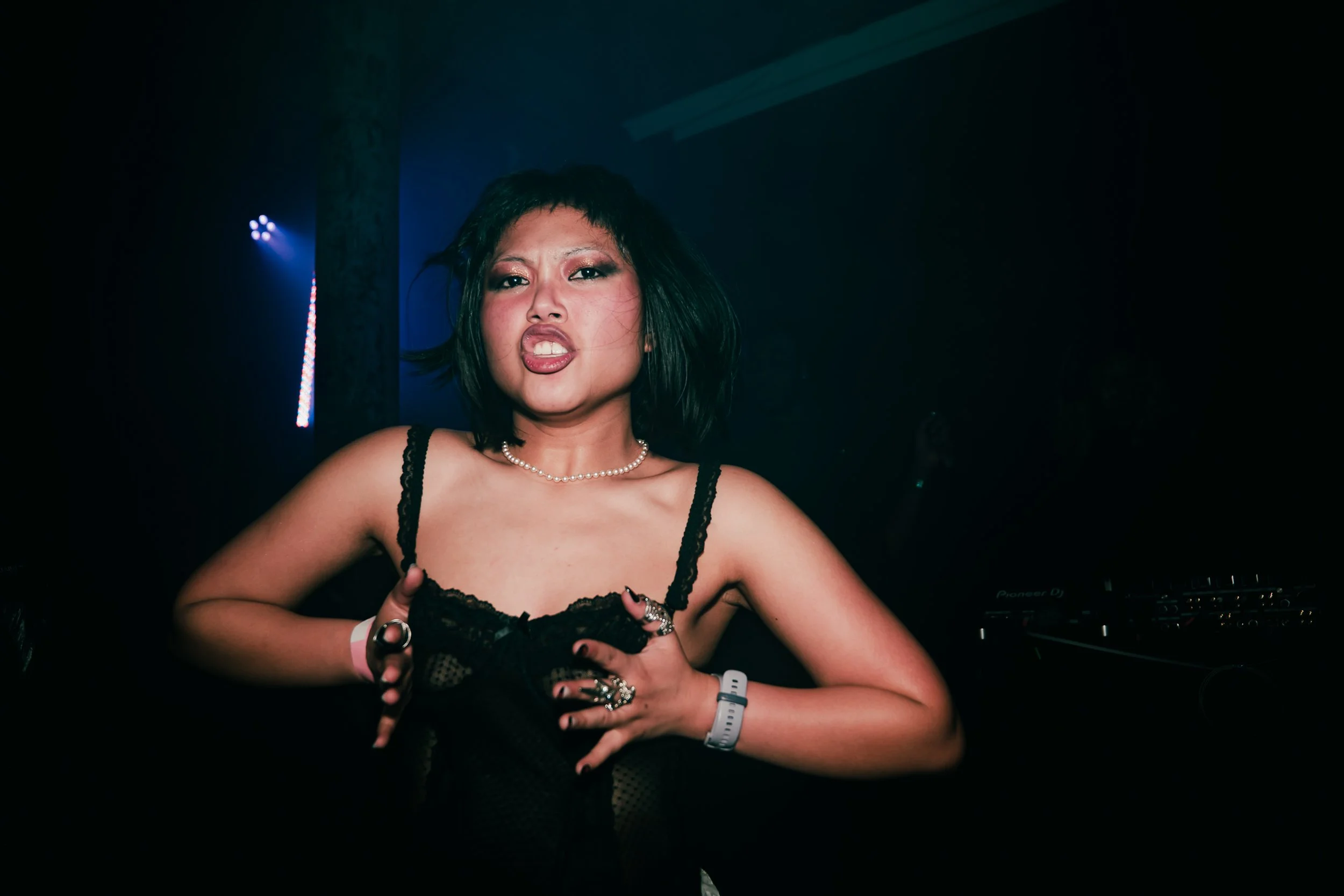 Woman with black hair, makeup, and jewelry, wearing a black lace top, in a dark club or concert setting with colorful lights around her.