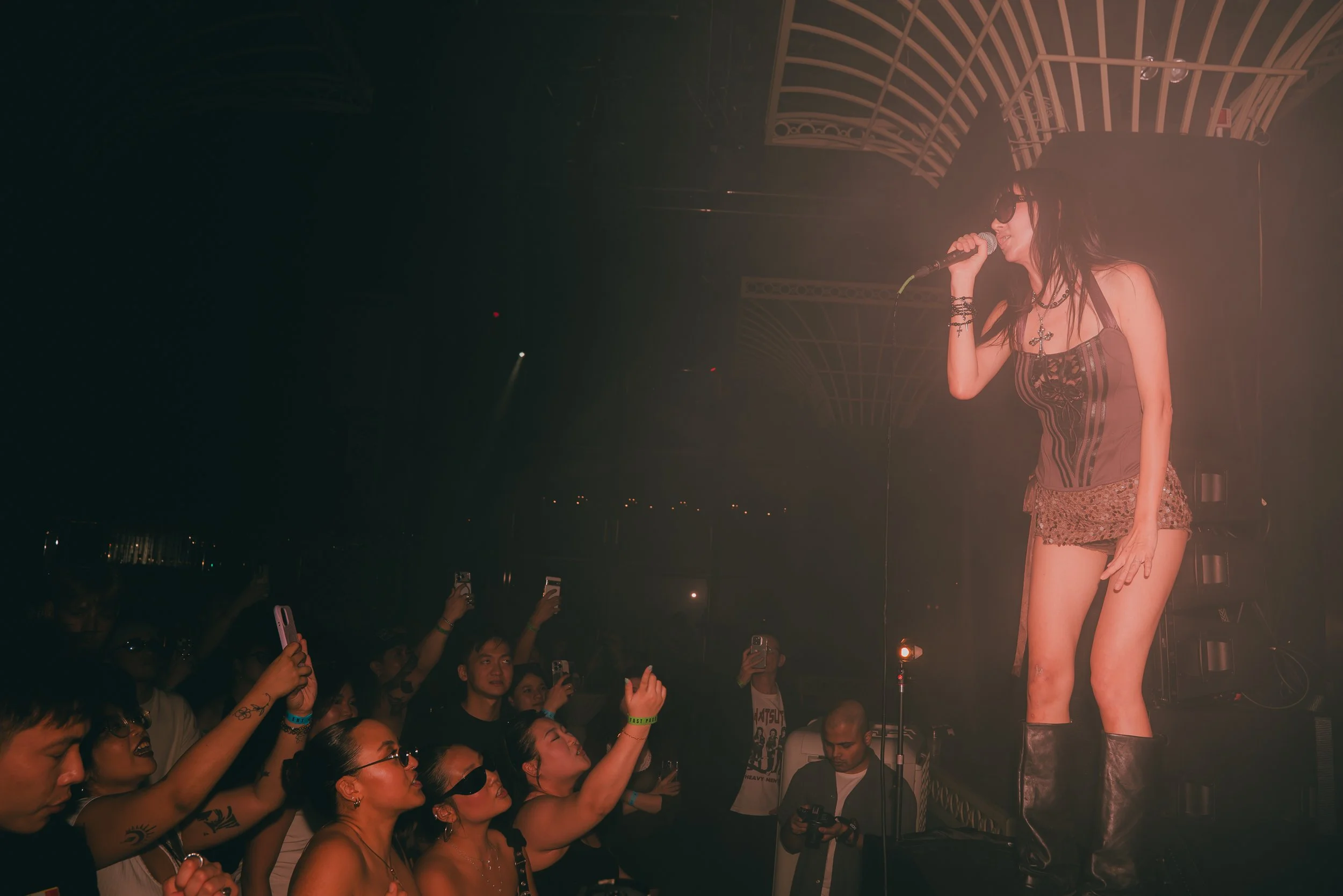 A singer wearing sunglasses, a harness-style top, and leopard print shorts performs on stage at night, surrounded by an audience taking photos and videos.