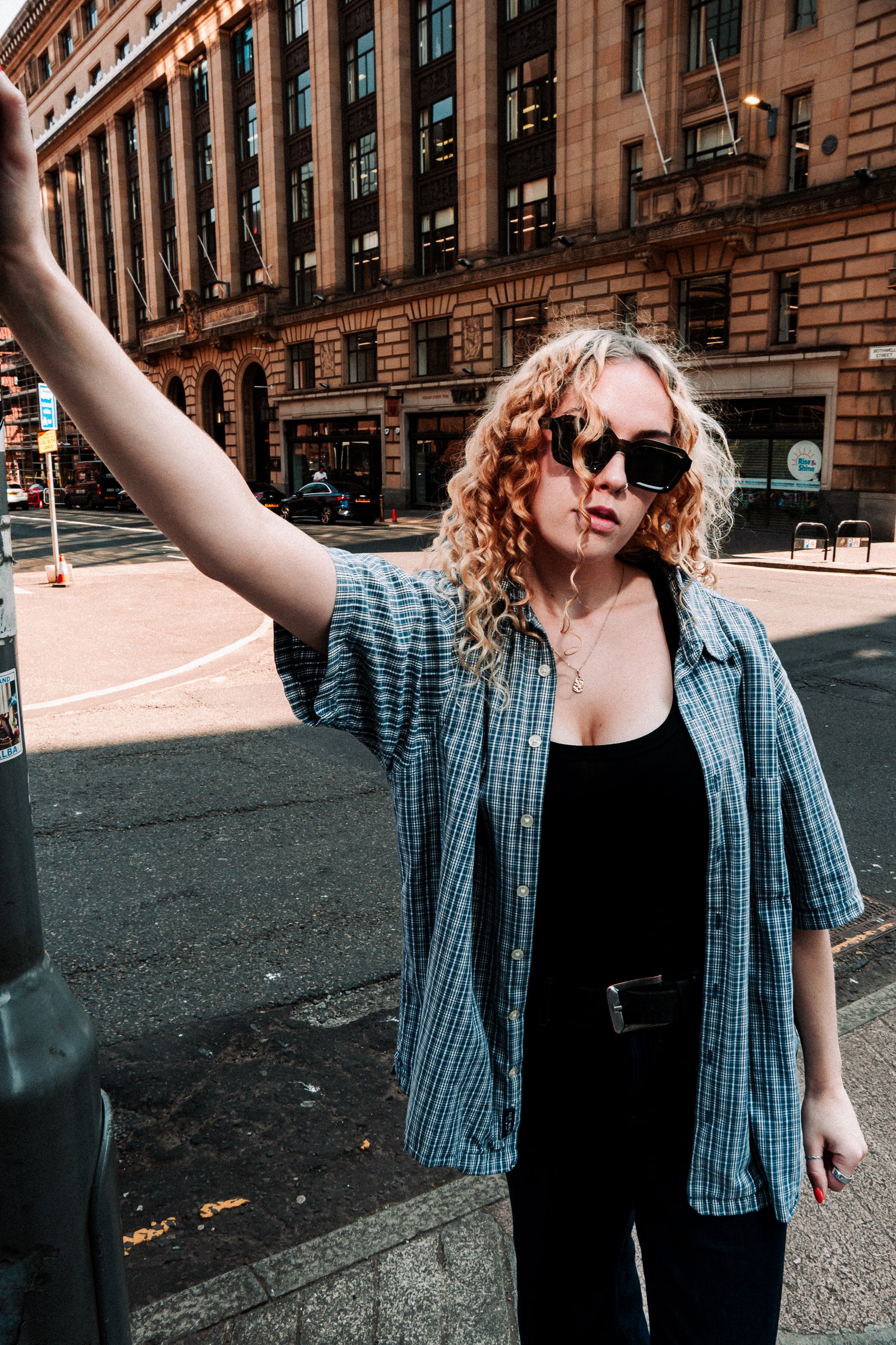 A woman with curly blonde hair wearing dark sunglasses, a black top, and a blue plaid shirt, standing on a city street with a large brick building in the background.