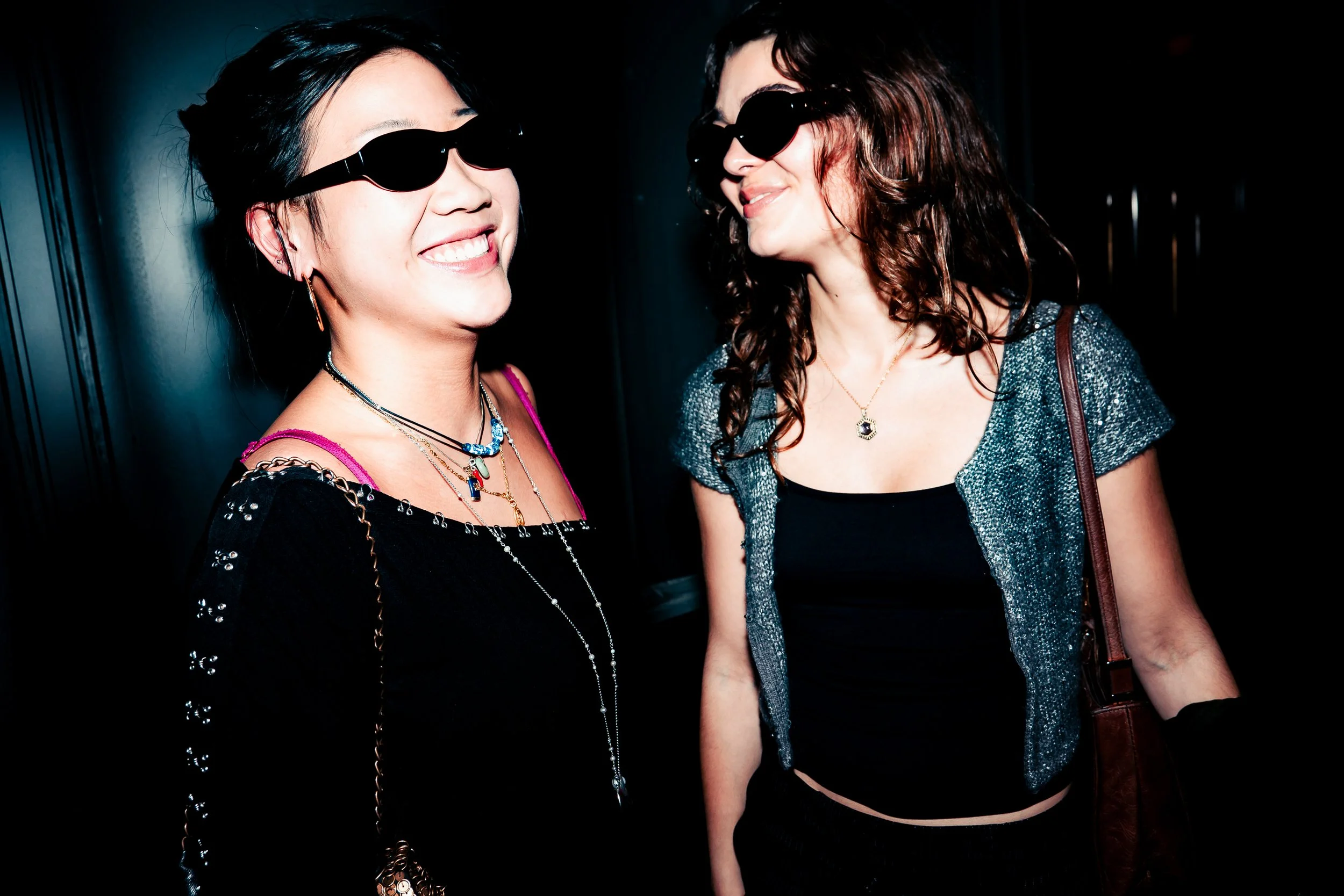 Two women wearing black sunglasses and smiling, engaged in conversation, in a dark indoor setting.