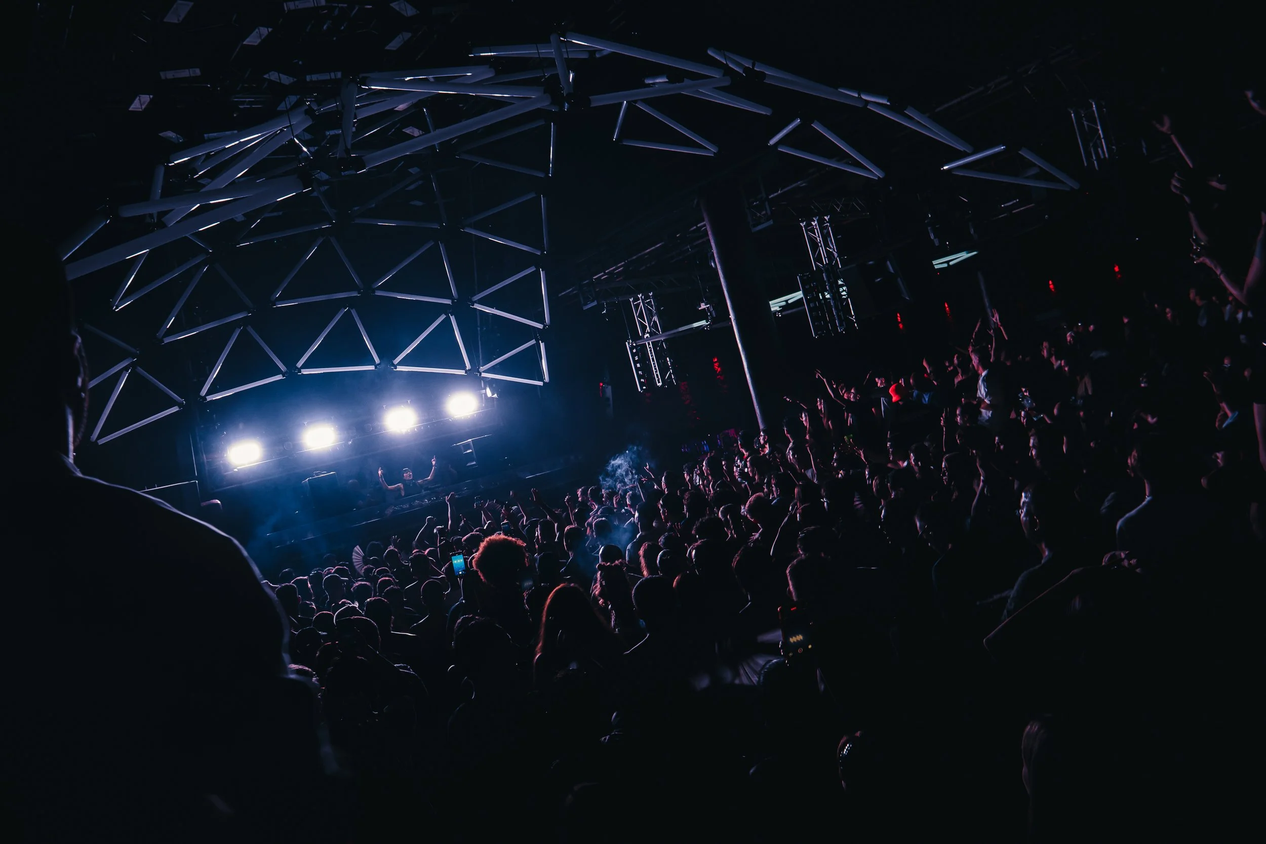 A large crowd of people at a concert or nightclub, dancing under bright stage lights and a modern geometric ceiling structure with illuminated beams.