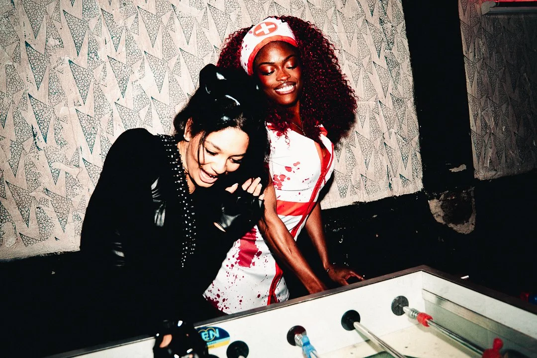 Two women dressed as a nurse and a nurse zombie playing table hockey together, smiling.