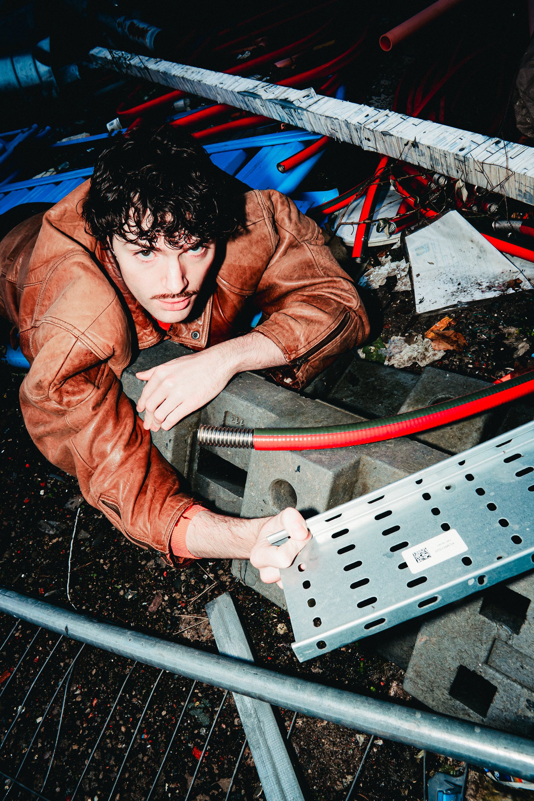 A man with dark curly hair, wearing a brown leather jacket, is working on a metal construction or repair project outdoors. He is laying on the ground and holding a screwdriver near a metal perforated panel. The background features various metal and p