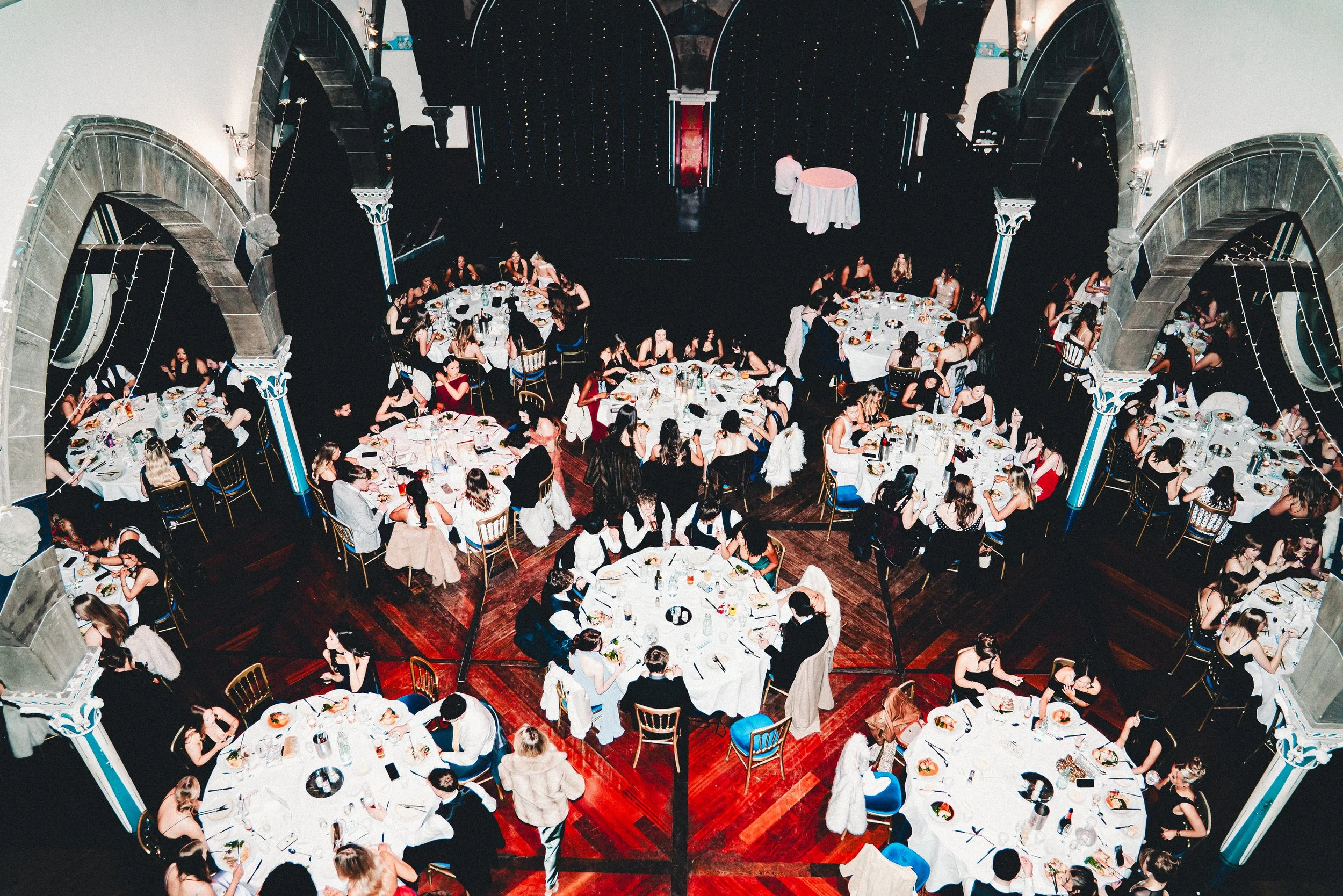 A large banquet hall filled with oval tables covered in white tablecloths, each with multiple guests dining and socializing. The hall has ornate stone arches and decorative columns. Small lights hang from the ceiling, creating a festive atmosphere. O