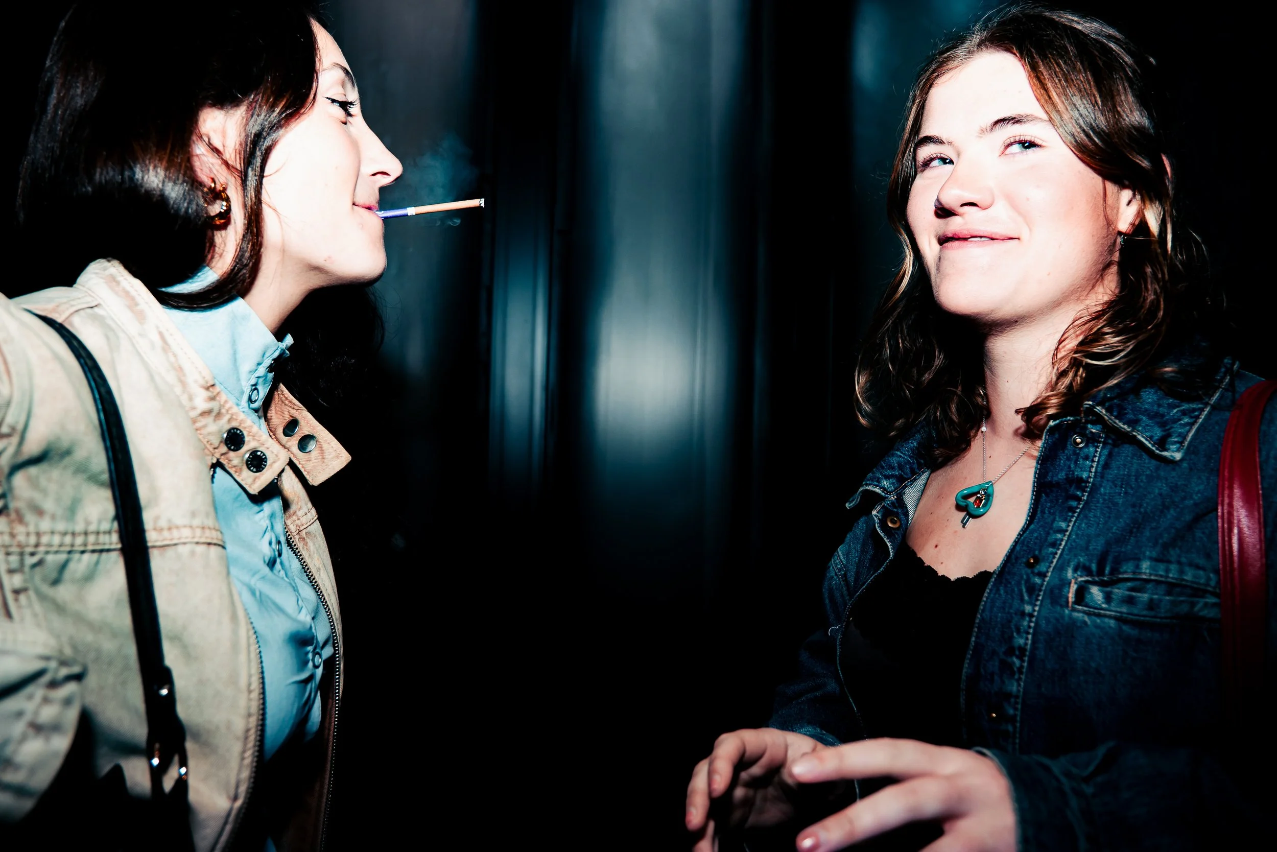 Two young women standing close in an elevator, one smoking and the other looking at the camera. Both are dressed casually, one with a denim jacket and the other with a beige jacket, with a dark background.