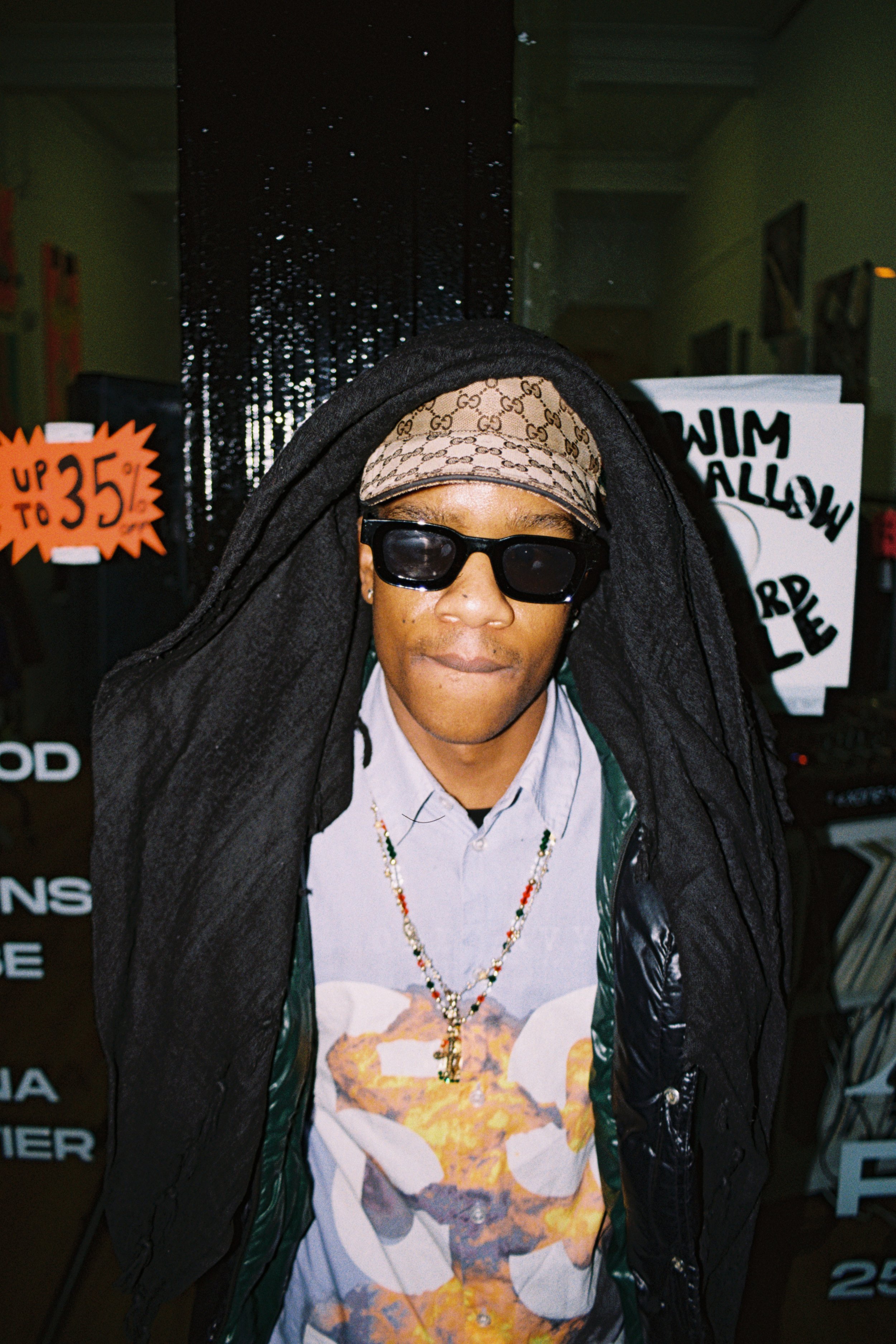 Person wearing sunglasses, a Gucci headband, a black hoodie, a tie-dye T-shirt with a lion design, and several necklaces, standing indoors with various signs and posters in the background.
